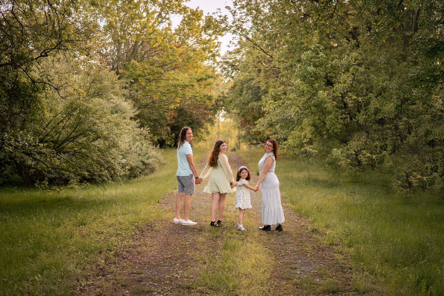 A group in white dresses stands on a sunlit path surrounded by lush green trees during golden hour.
