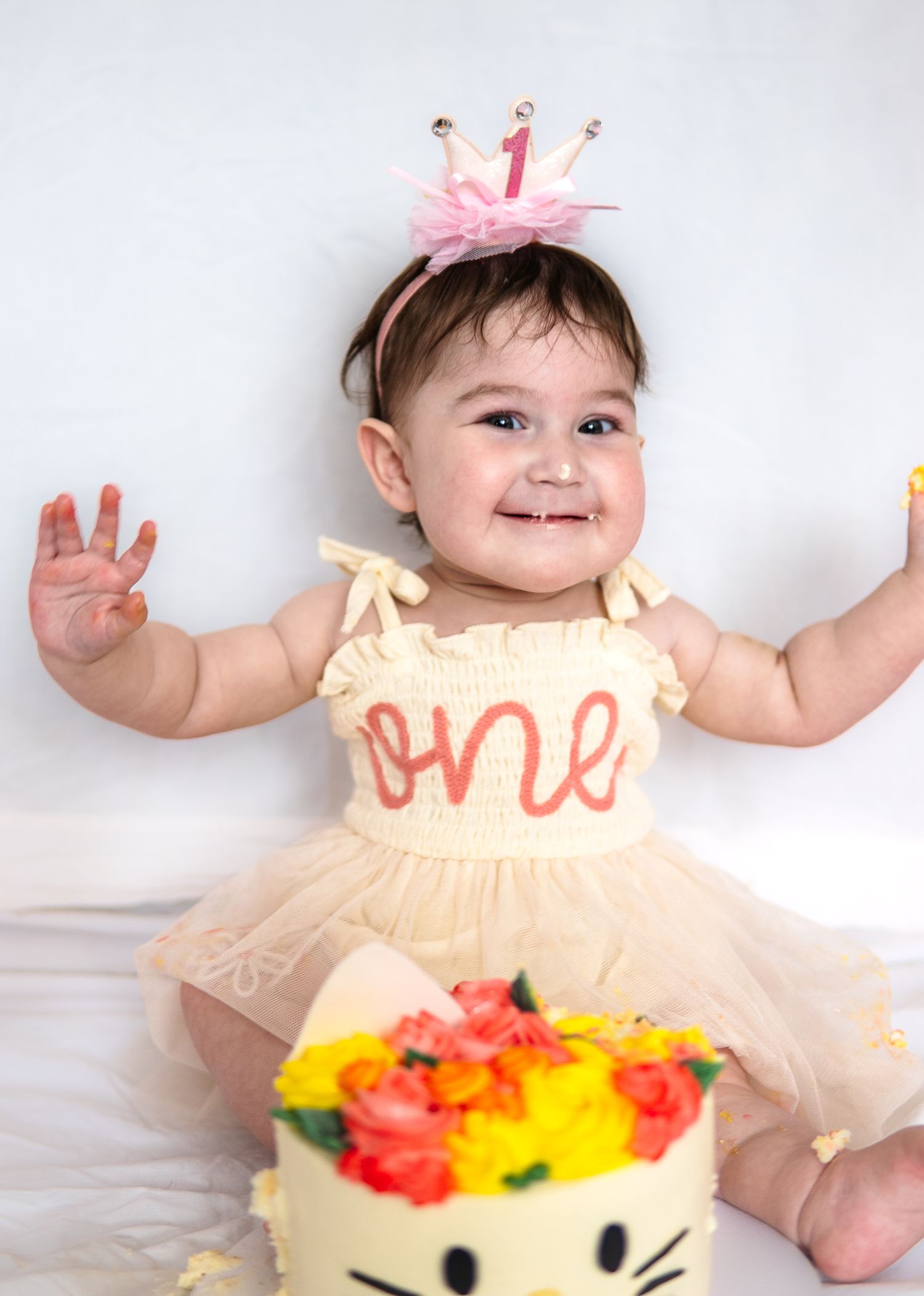 Excited baby in crown and 'one' dress poses with floral decorated Hello Kitty birthday cake against white backdrop.
