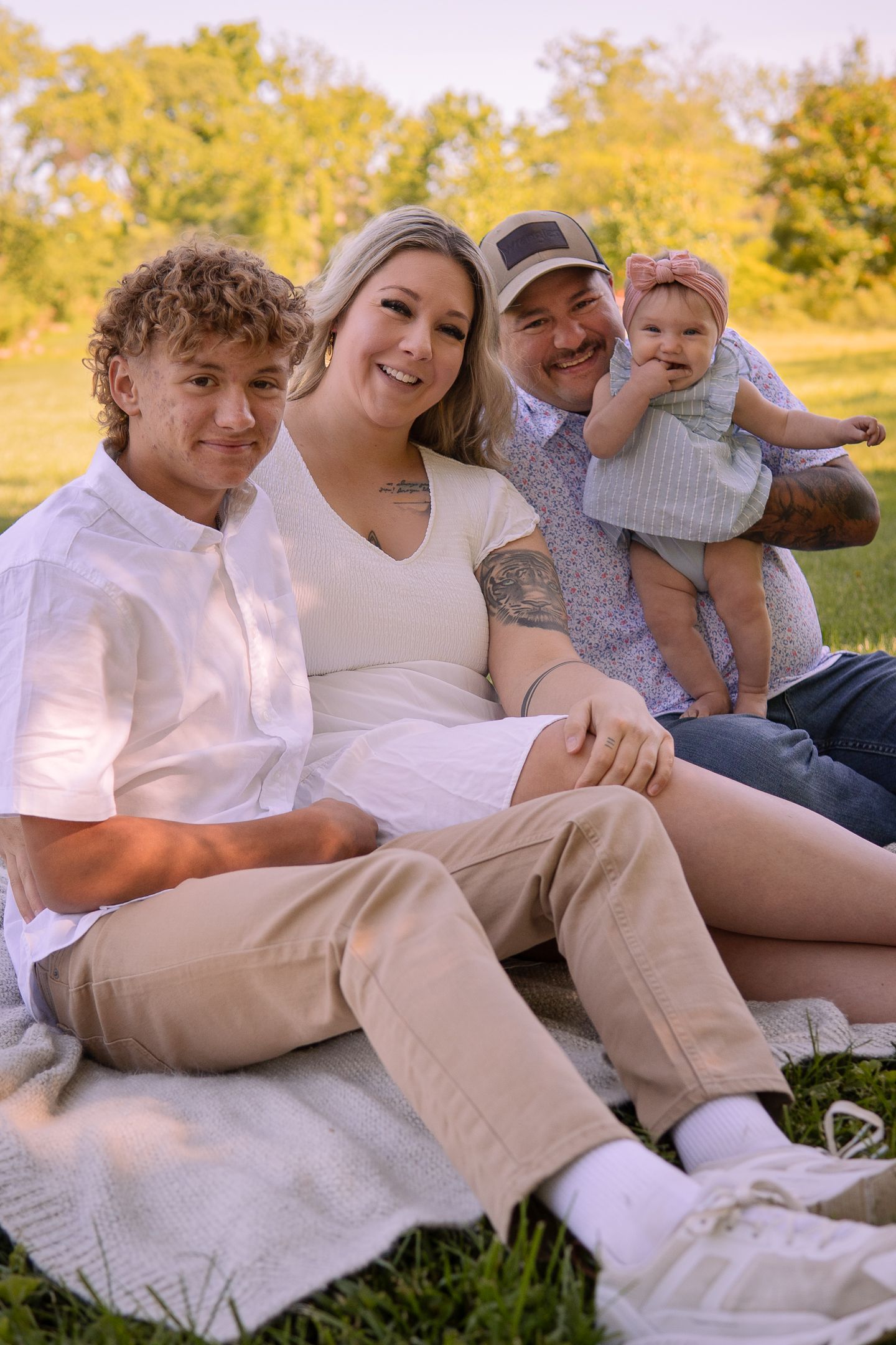 A family relaxes together on a blanket in a sunny park with golden autumn trees in the background.