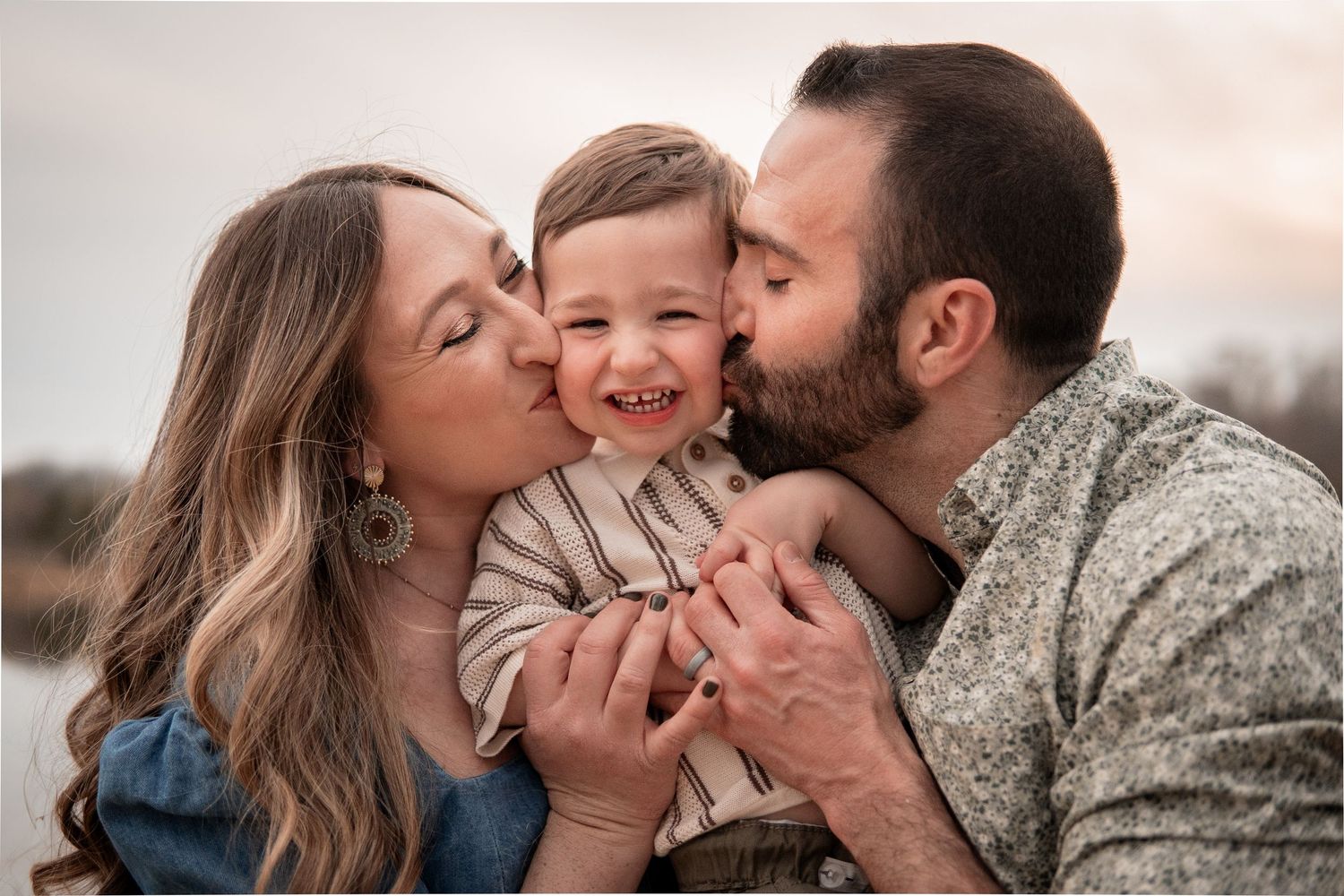 Happy family sharing kisses and laughter together during an outdoor portrait session at sunset.