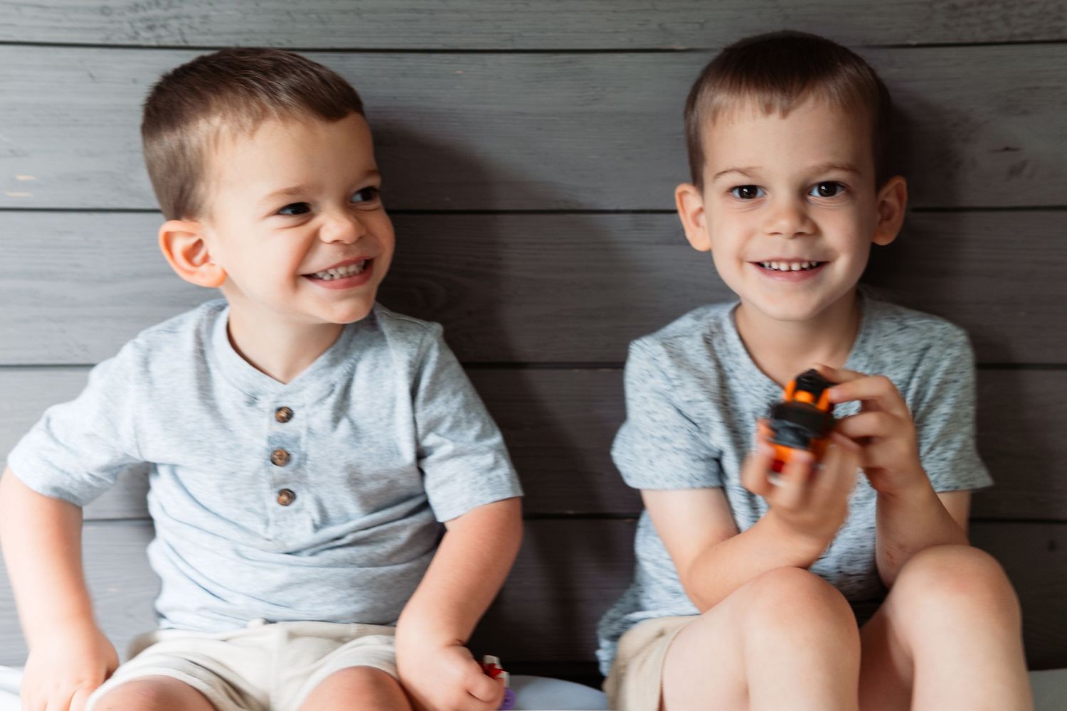 Young boy in light blue shirt sitting and smiling against grey wooden wall.