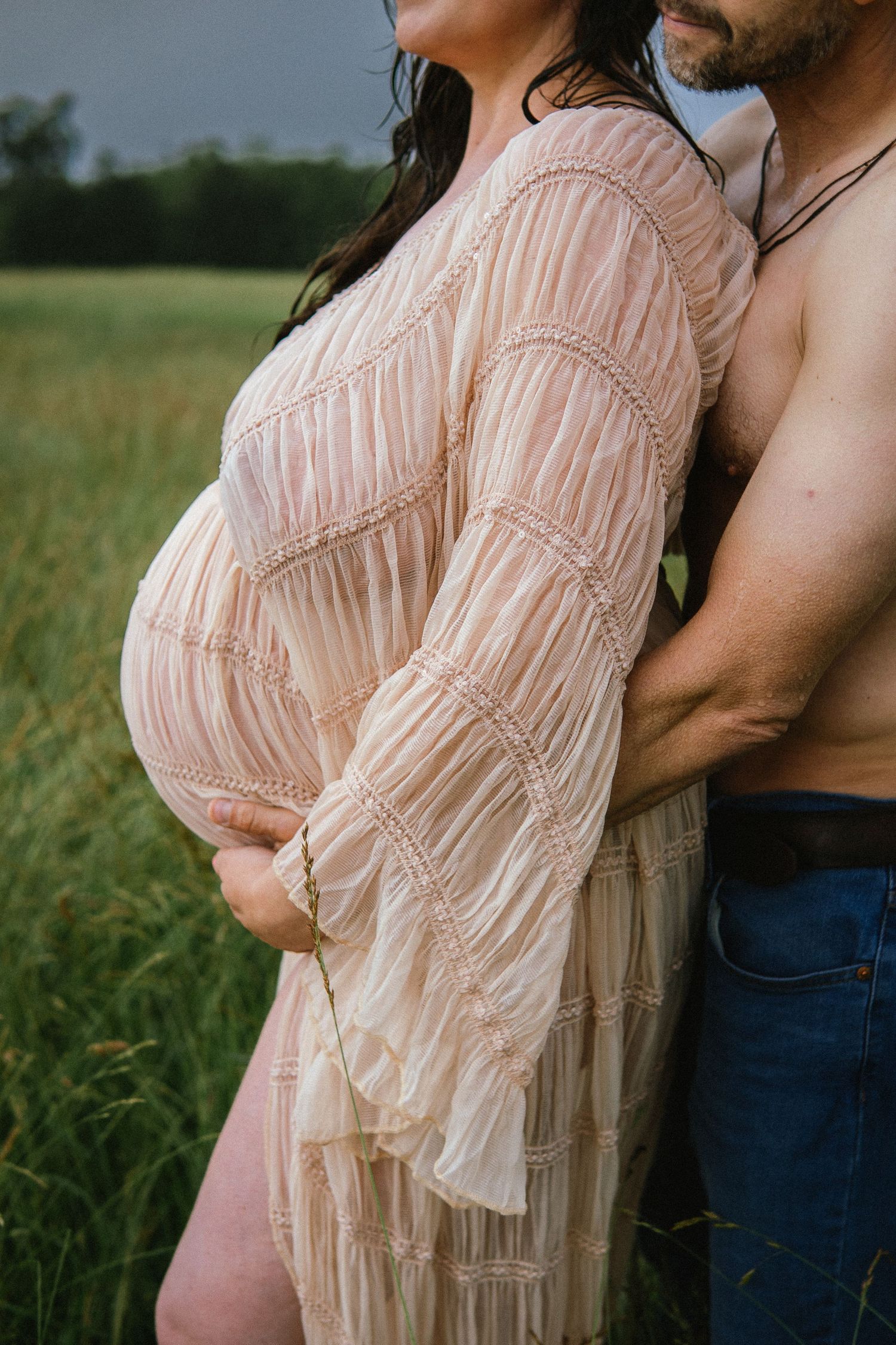 A pregnant figure wearing a romantic blush-colored lace dress stands close to their partner in a grassy field.
