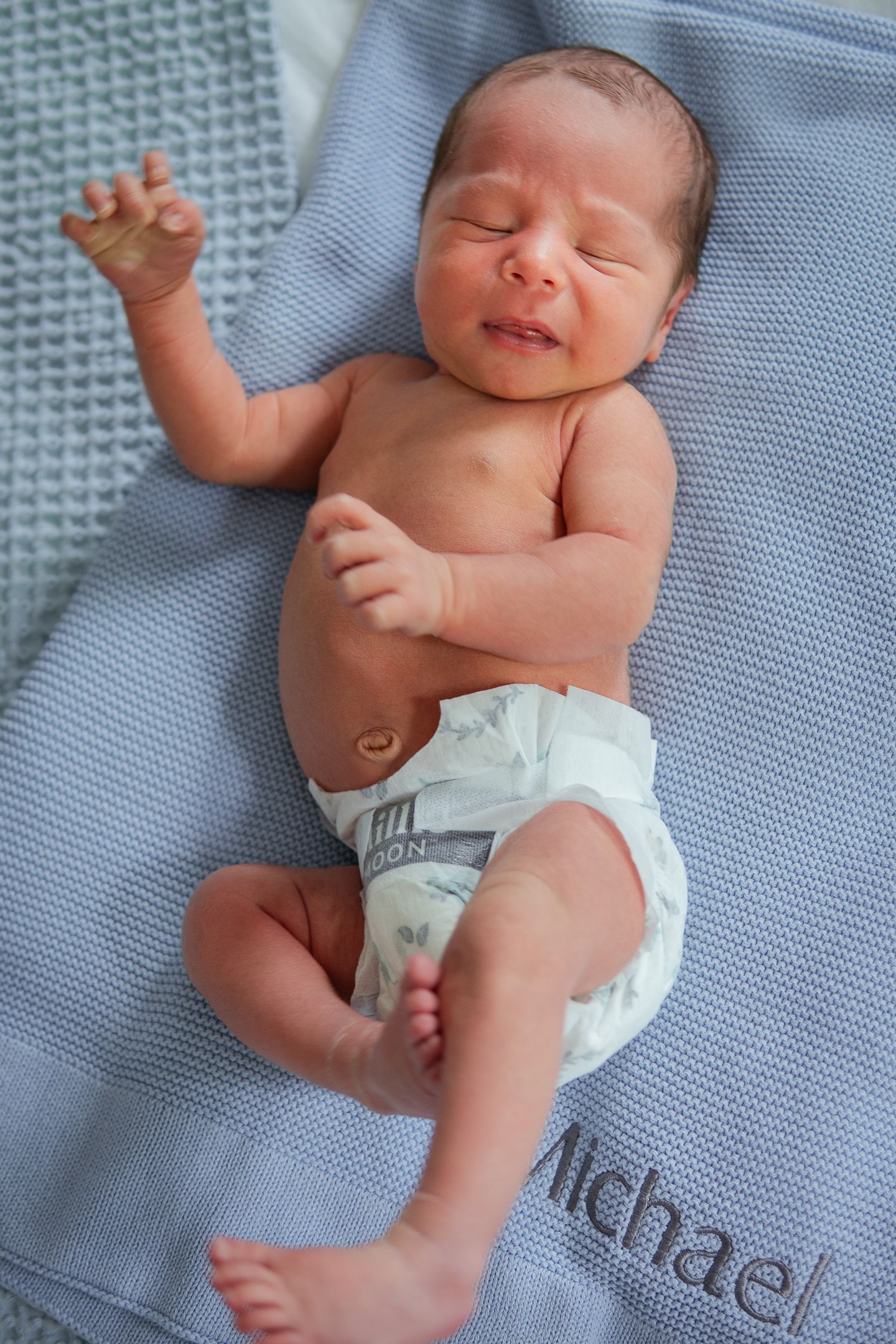 Newborn baby wearing diaper stretches while resting on blue knitted blanket.