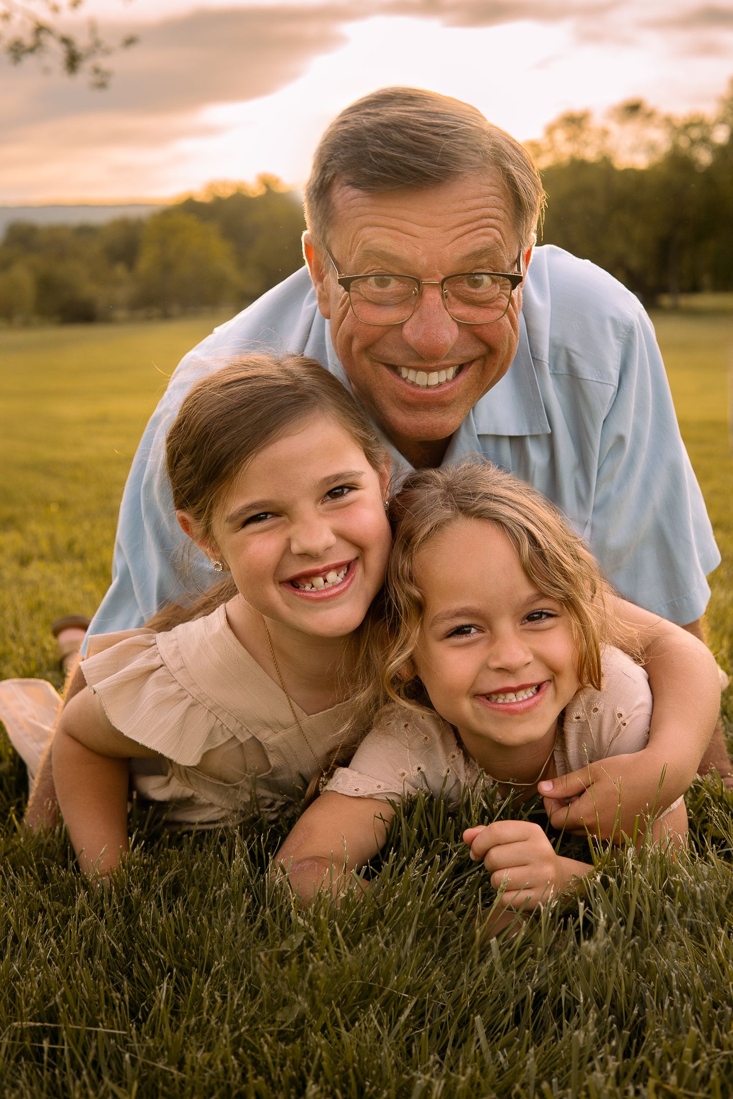 Three people laying in grass with bright smiles at sunset, wearing casual clothes in a pastoral outdoor setting.