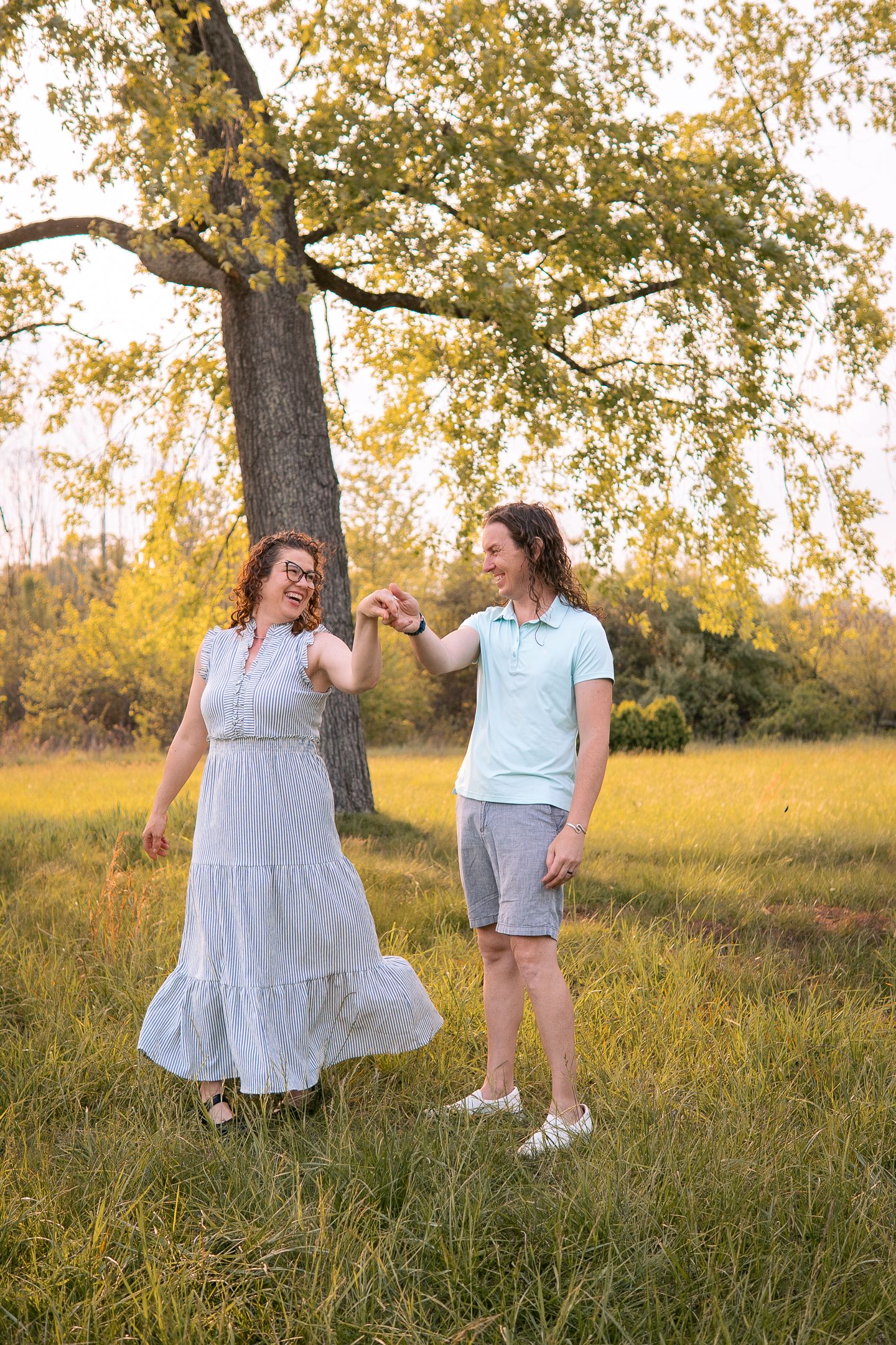 A couple stands together in a sunny meadow beneath a large tree with golden autumn leaves and tall grass.