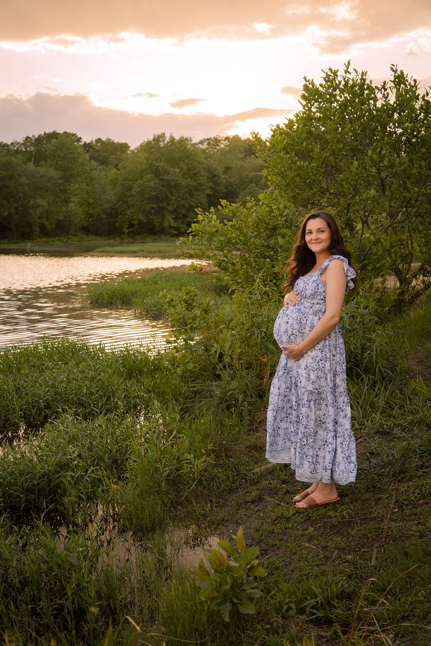 A woman in a floral dress stands by a serene lake at sunset among green foliage.