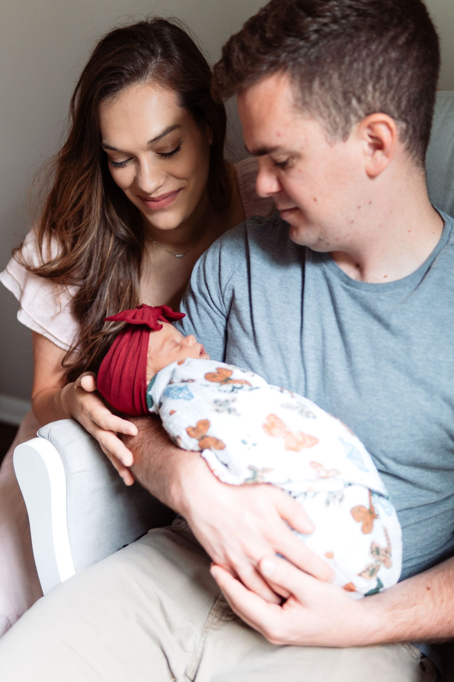 Parents cradle their newborn baby while sitting together on white furniture in natural lighting.
