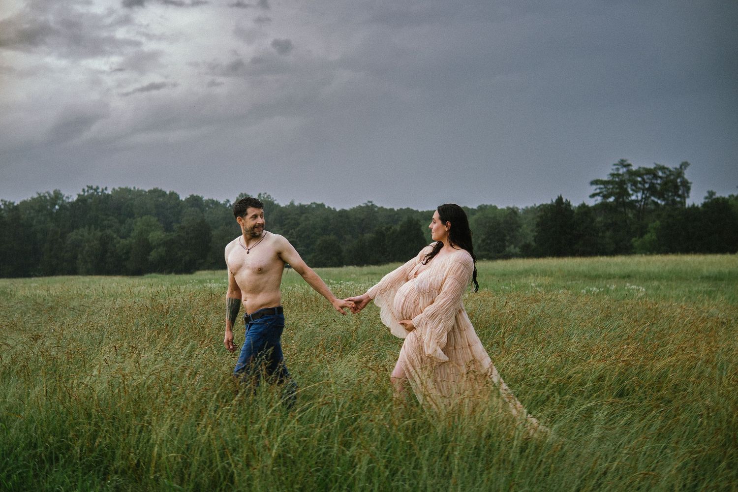 Two people hold hands while walking through a misty green field at sunset wearing romantic coordinated outfits.