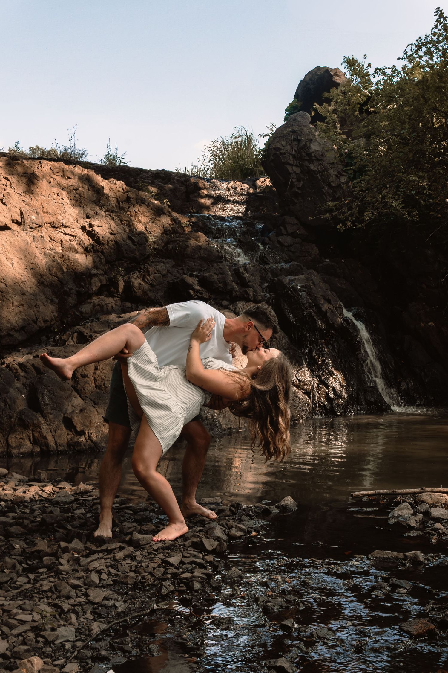 A dancer performs a graceful pose on rocky terrain near a waterfall in a dramatic nature photoshoot.