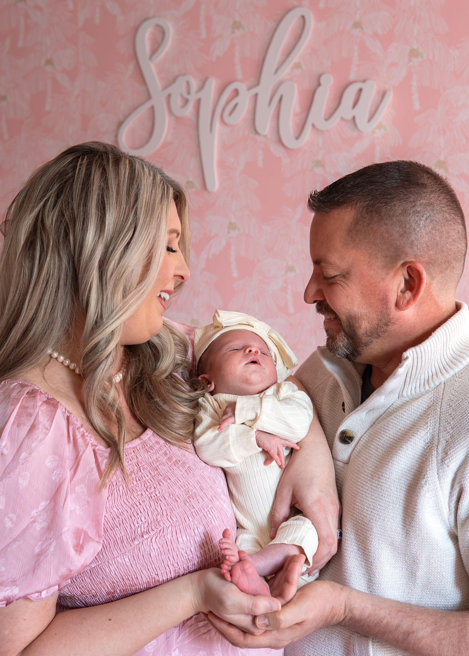 A tender family moment captured against a pink textured wall as parents lovingly gaze at their newborn baby.