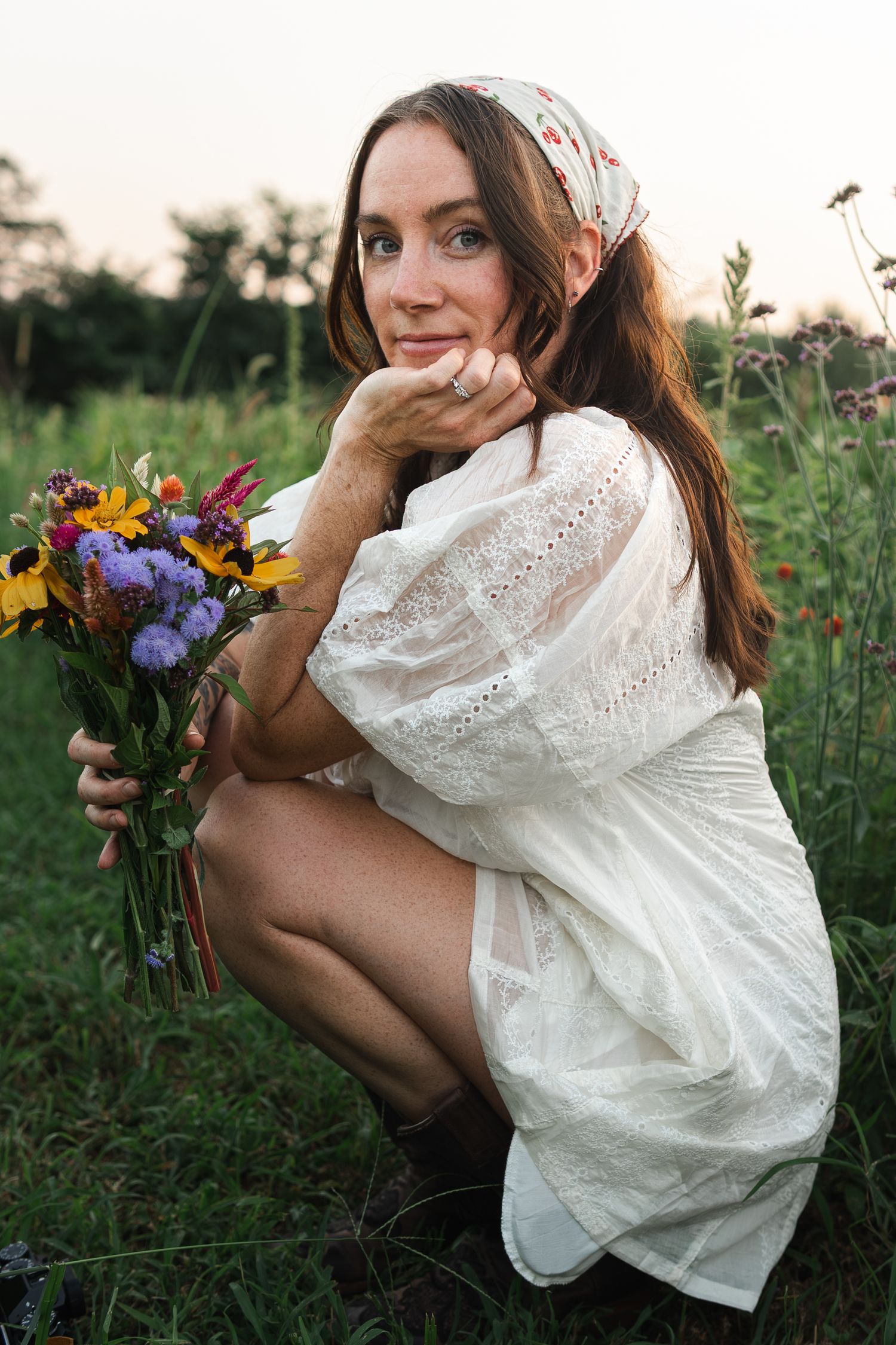 Woman in white lace dress crouches in meadow at sunset holding colorful wildflower bouquet.