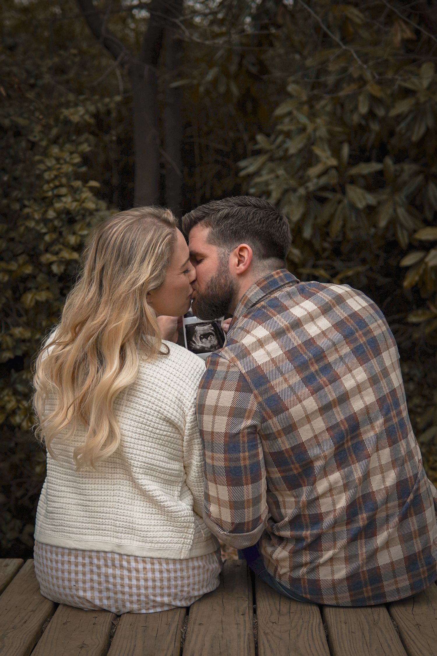 Romantic couple in casual autumn attire share an intimate moment while sitting on wooden deck surrounded by fall foliage.