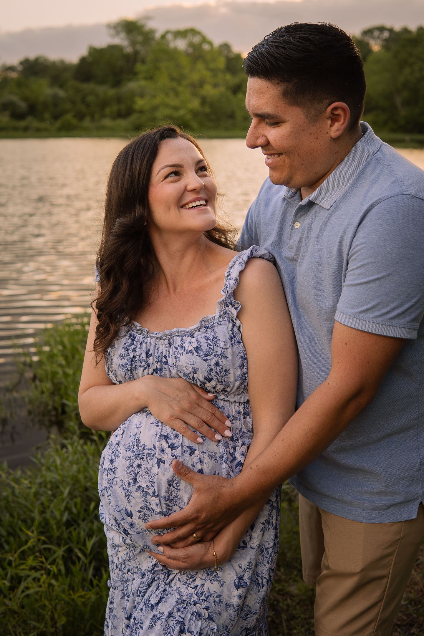 A couple embraces by a lake at sunset while wearing a blue floral dress and grey polo shirt.