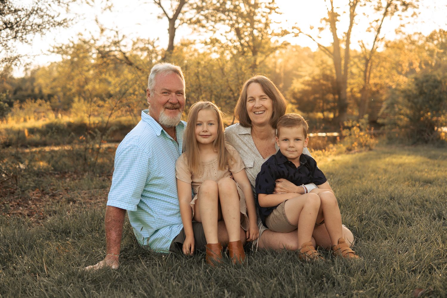 A family of four sits together on grass during golden hour sunset in a natural outdoor setting in Haymarket, Virginia.