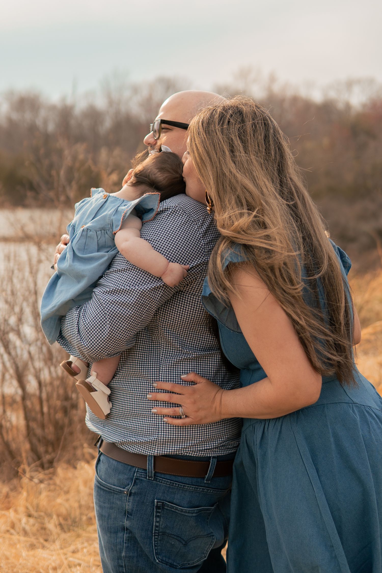Family wearing coordinating blue outfits embrace in a golden field during sunset.