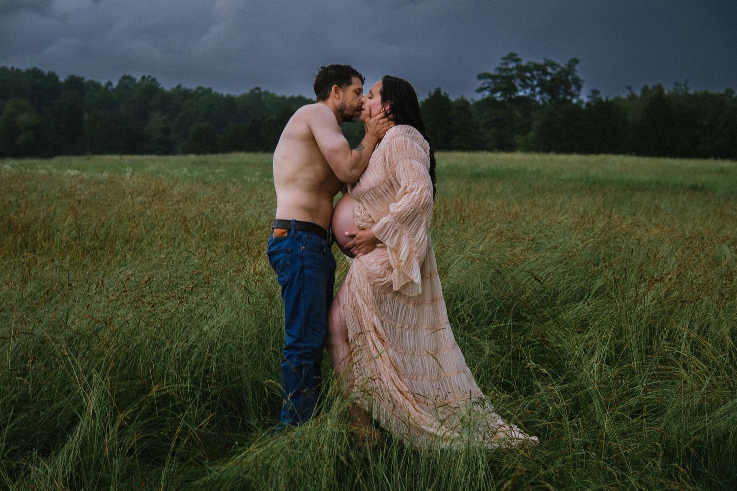 A couple shares an intimate moment in a meadow with dramatic storm clouds overhead.