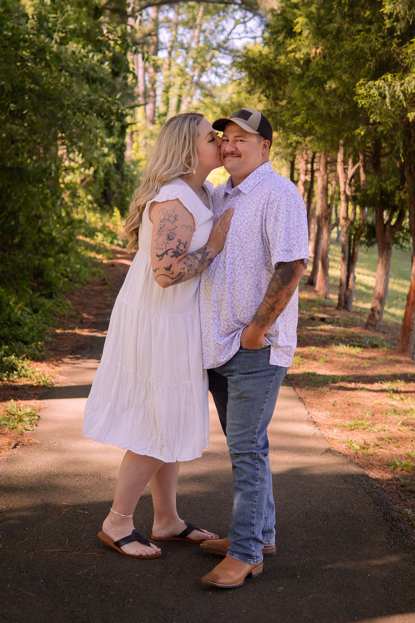 A couple sharing a tender moment on a tree-lined path, with the person in white wearing a flowing dress and visible tattoos.