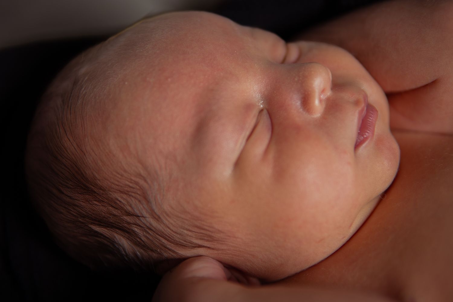 Close-up side profile of a sleeping newborn baby with delicate features in soft lighting.