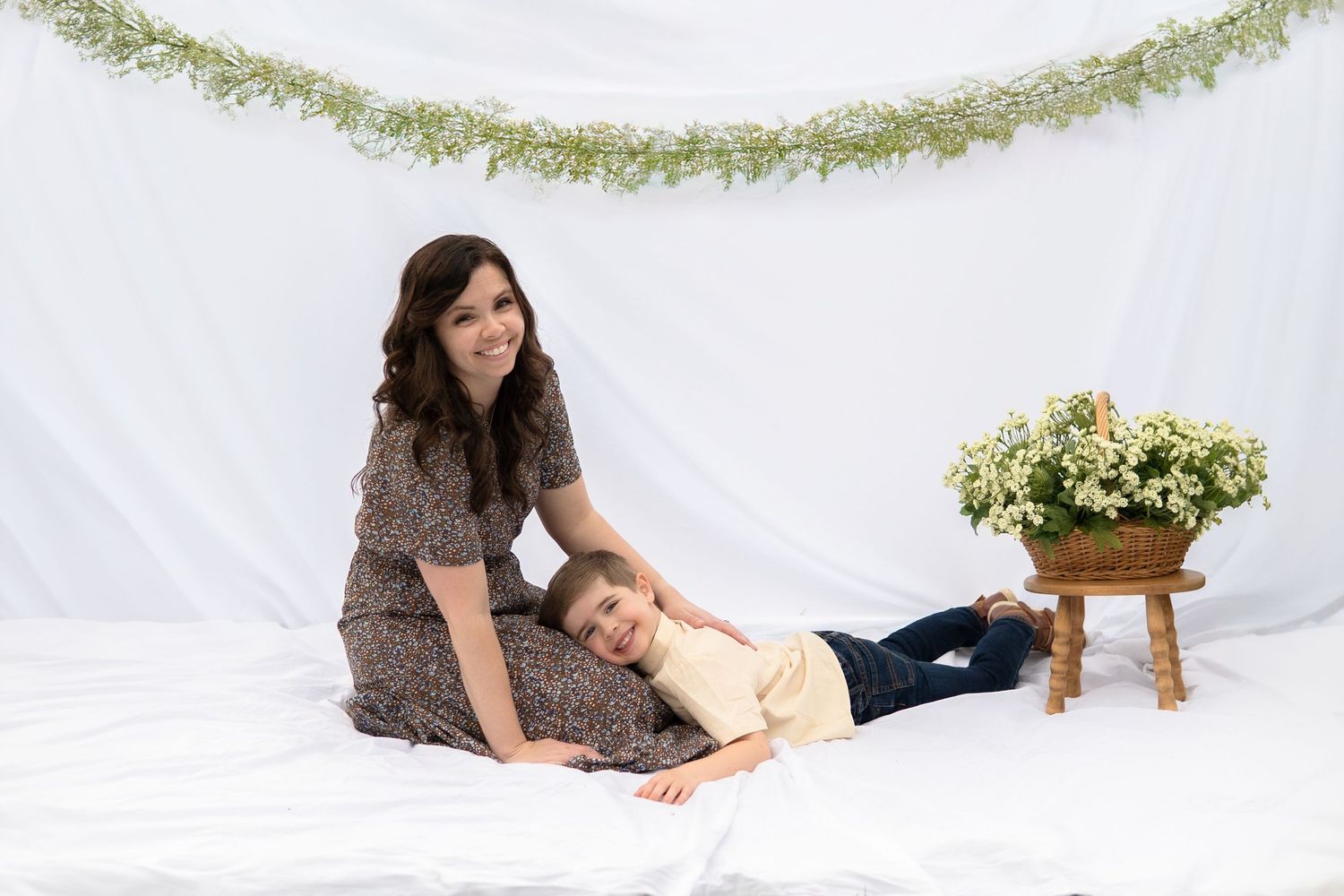 Two people pose together on a white backdrop with a green floral garland and potted yellow flowers.