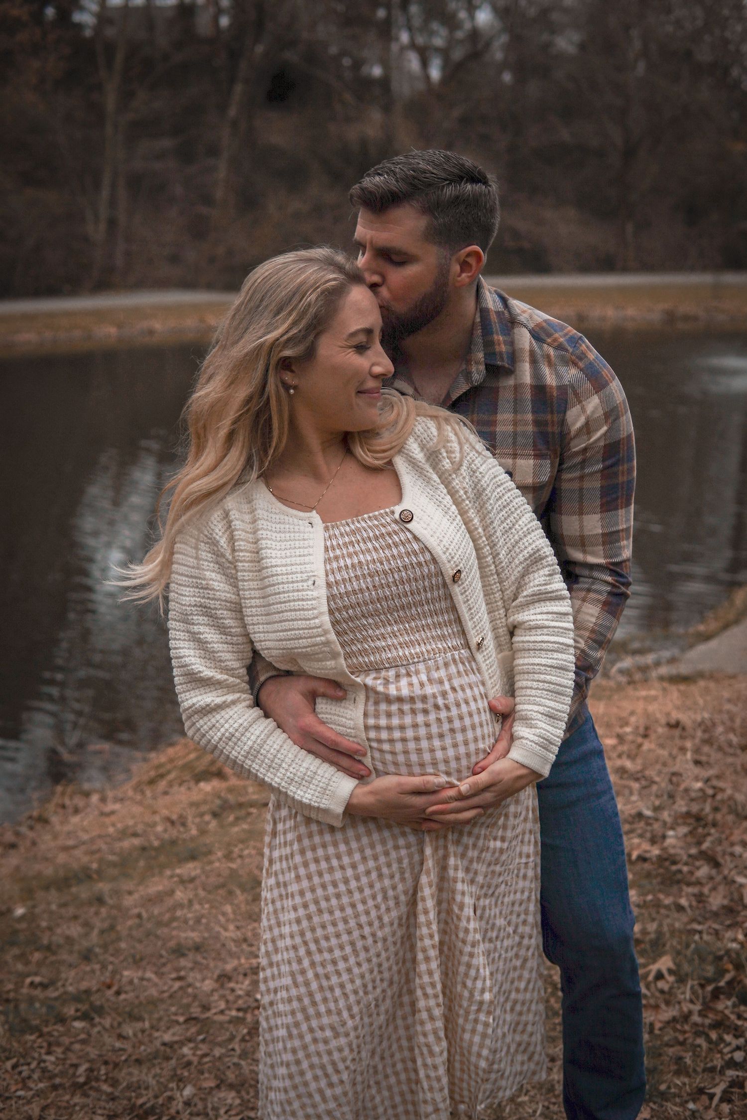 Expecting couple shares intimate moment by lake with her wearing cream dress and him in plaid shirt and jeans.