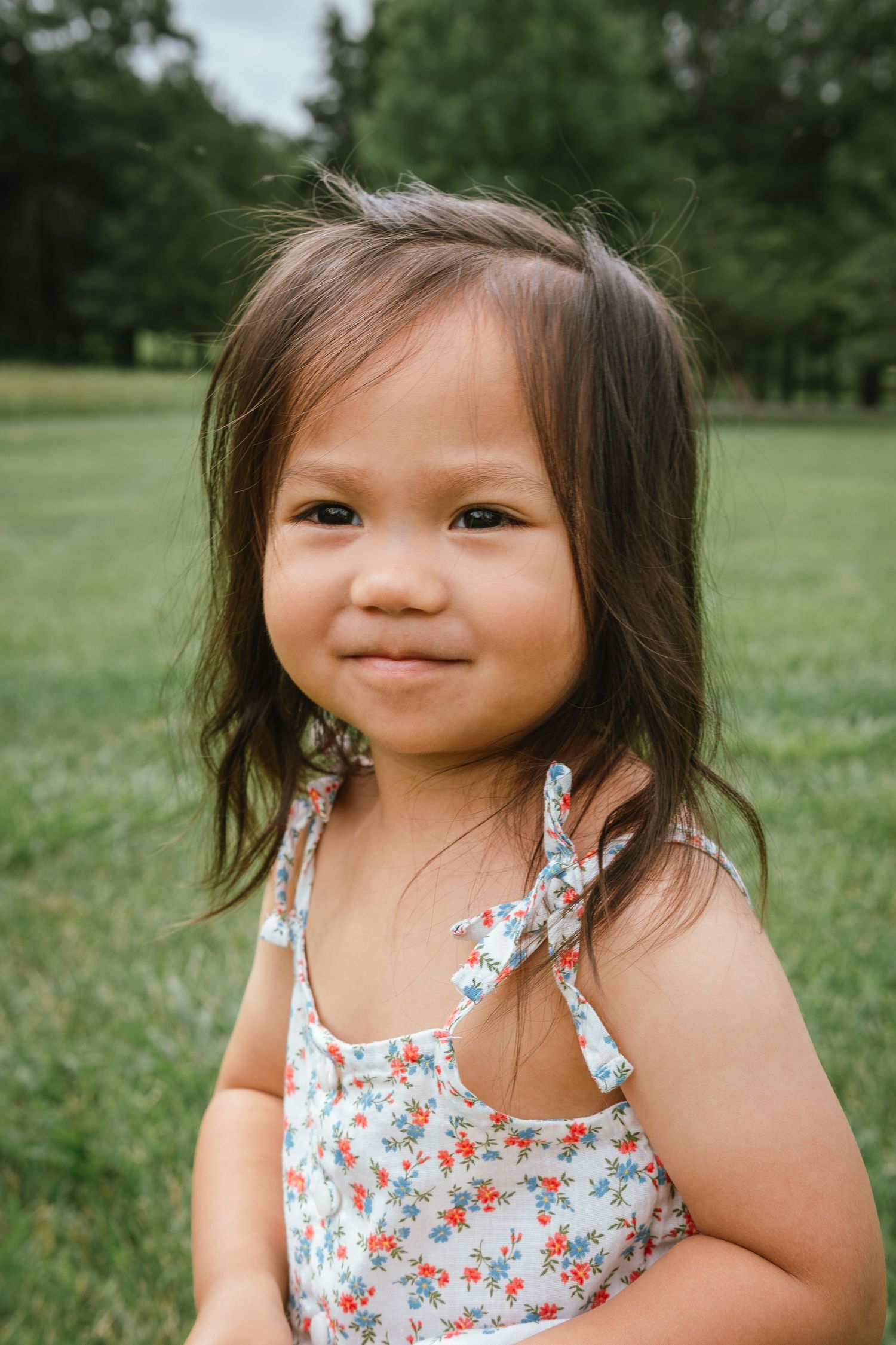 Young child in a floral sundress smiling brightly while playing outdoors in a grassy field.