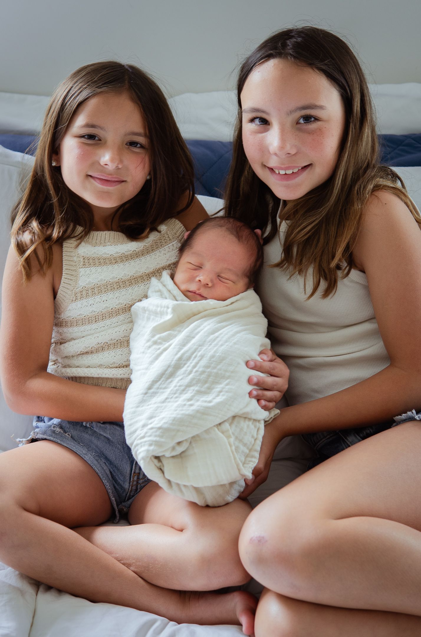 Two young girls sitting on bed and holding sleeping swaddled newborn between them.