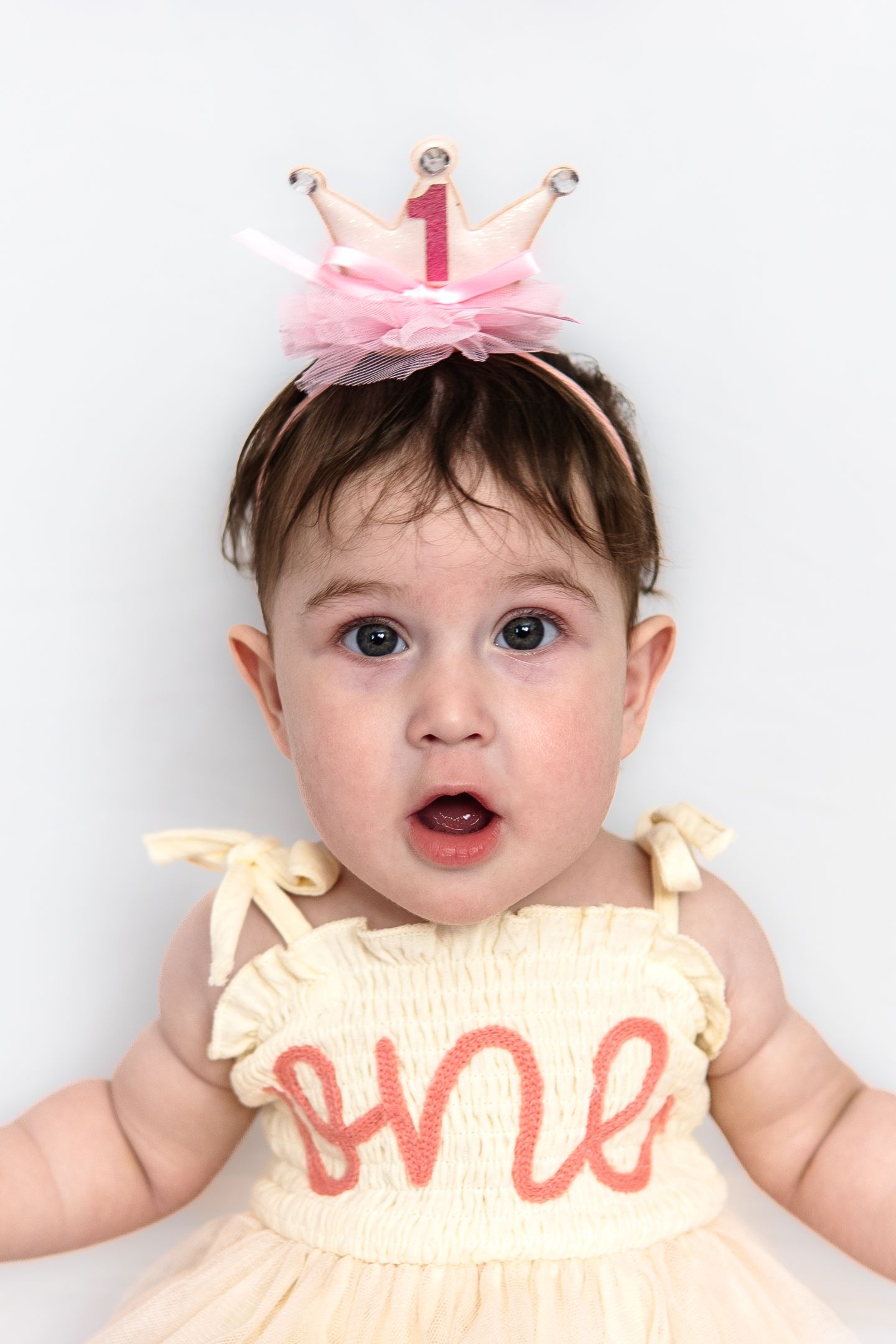 A smiling baby wearing a pink bow headband and cream-colored ruffled dress poses against a white background.