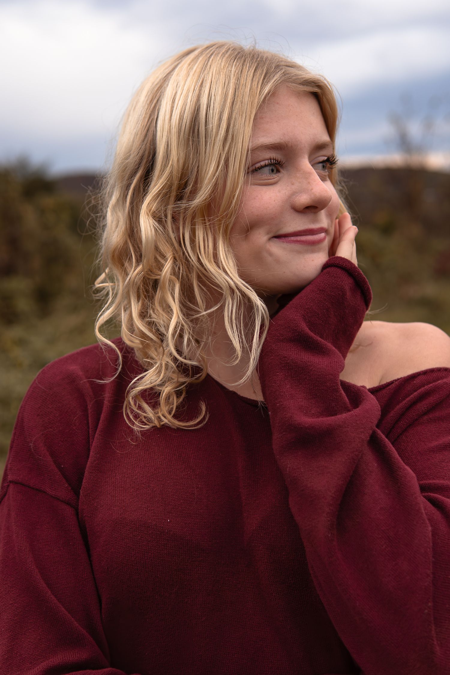 Person in maroon sweater smiles and poses against a natural outdoor background.