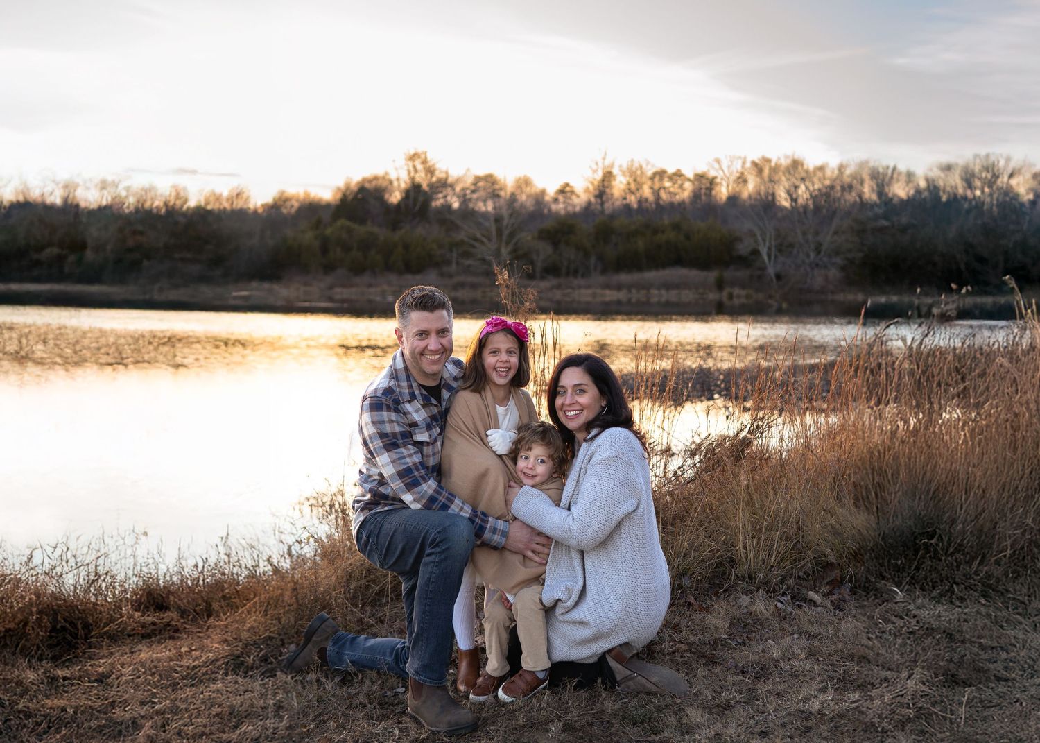 A couple poses together in a natural setting with tall grass and a lake at sunset.