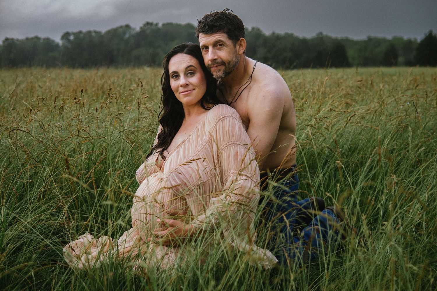 A couple embraces in a natural setting with tall grass and moody overcast skies in the background.