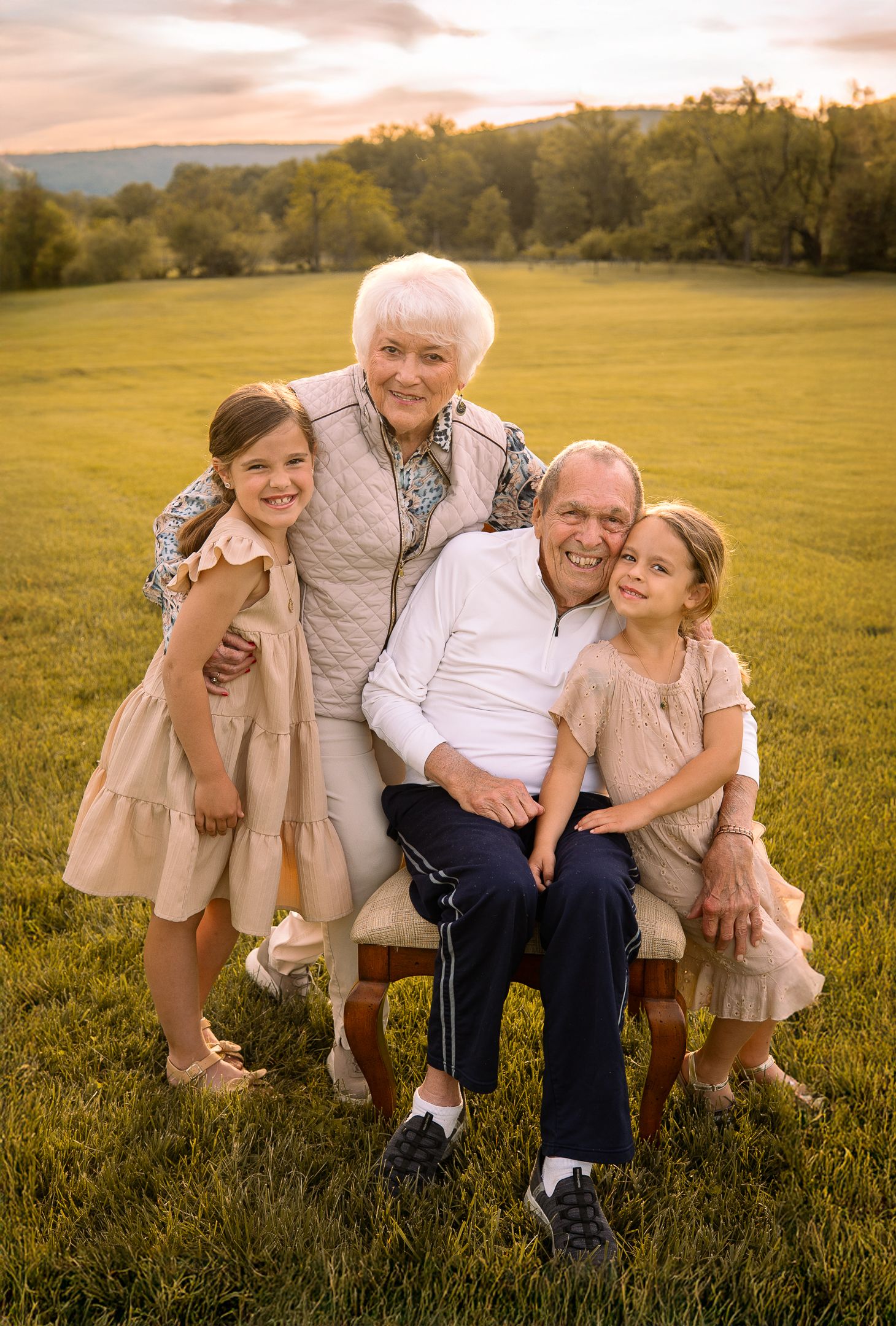 Multi-generational family portrait in a sunny meadow at sunset showing warm embraces and genuine smiles.