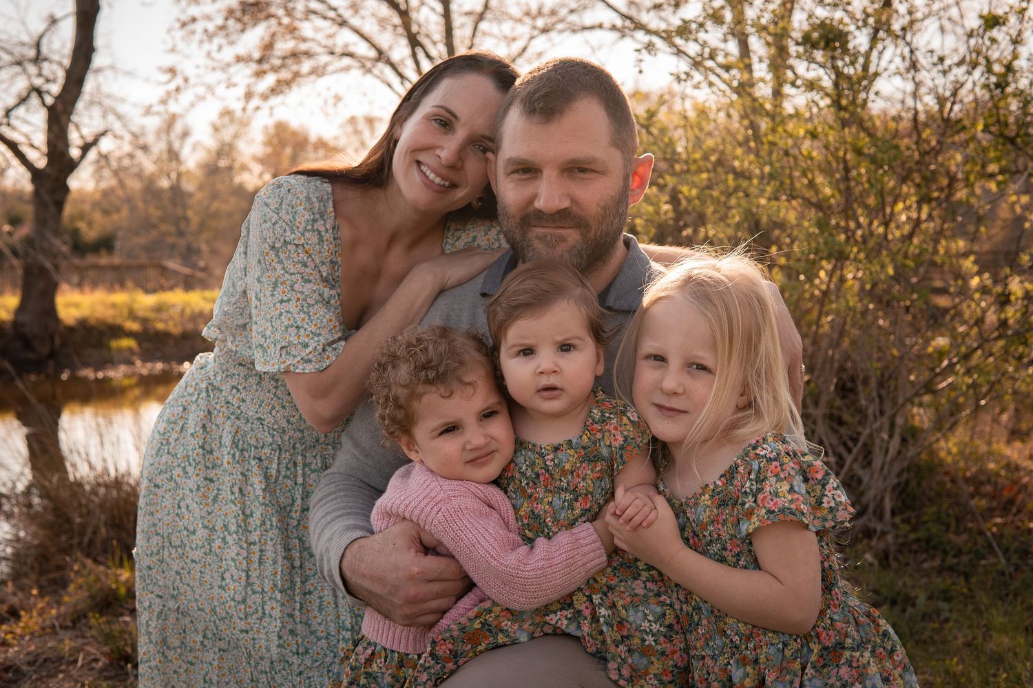 A family in coordinated floral outfits poses together outdoors during golden hour near a pond in autumn.