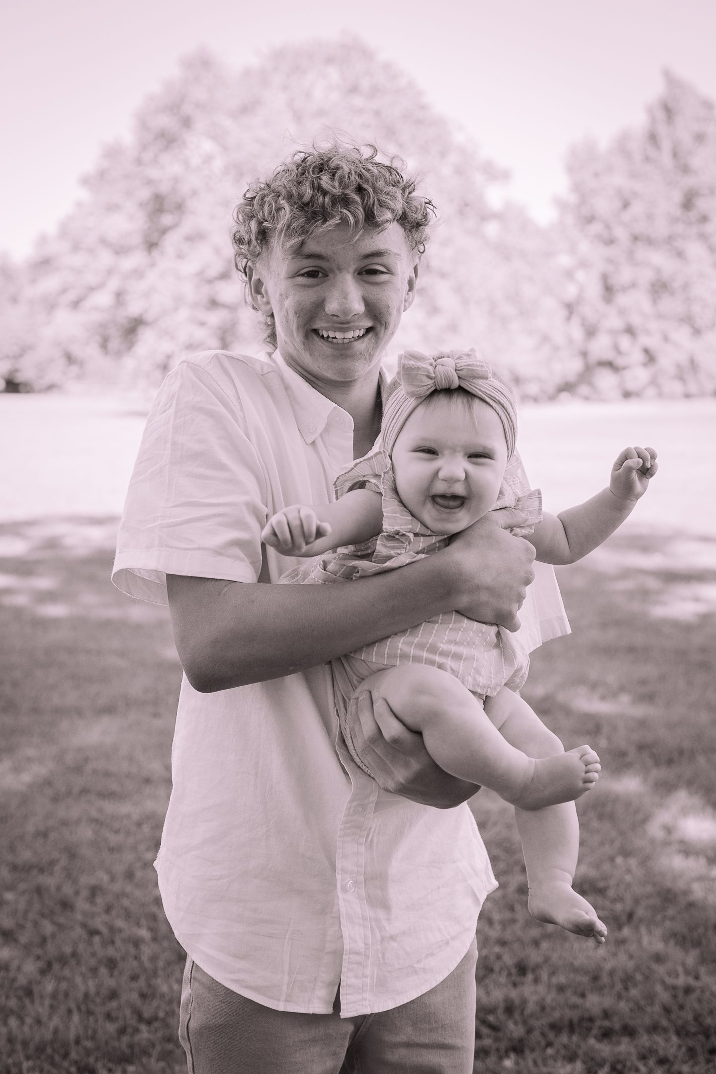 A black and white portrait of a teen holding a smiling baby outdoors on a sunny day.