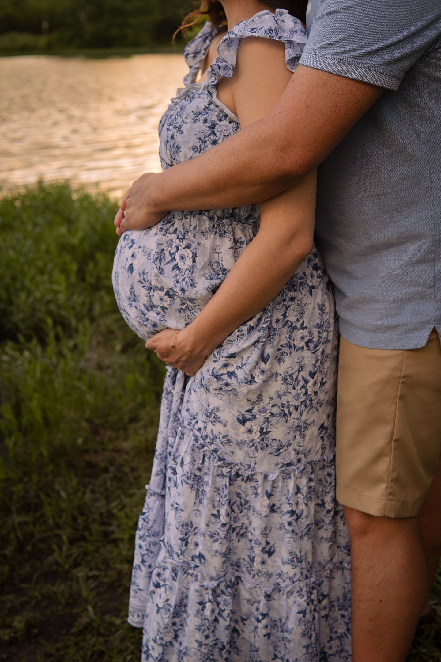 A pregnant woman in a blue and white floral dress stands beside someone wearing khaki shorts at sunset by a lake.