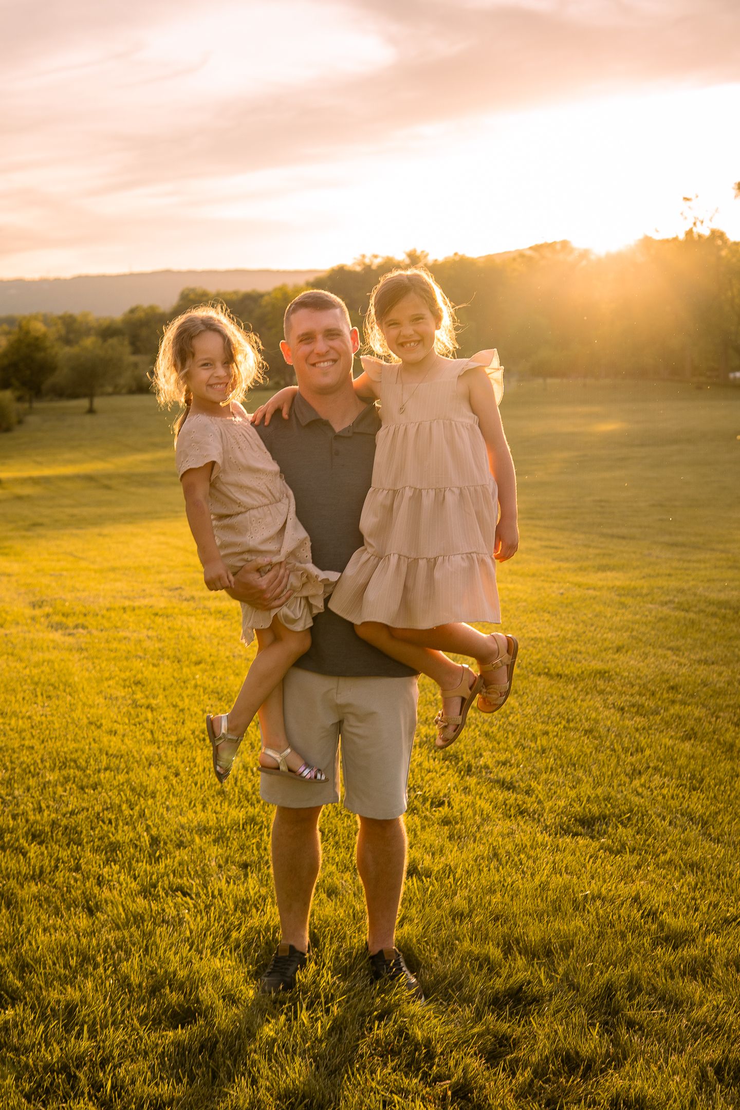 A warm sunset illuminates a grassy field as a person holds two young children in their arms during golden hour.