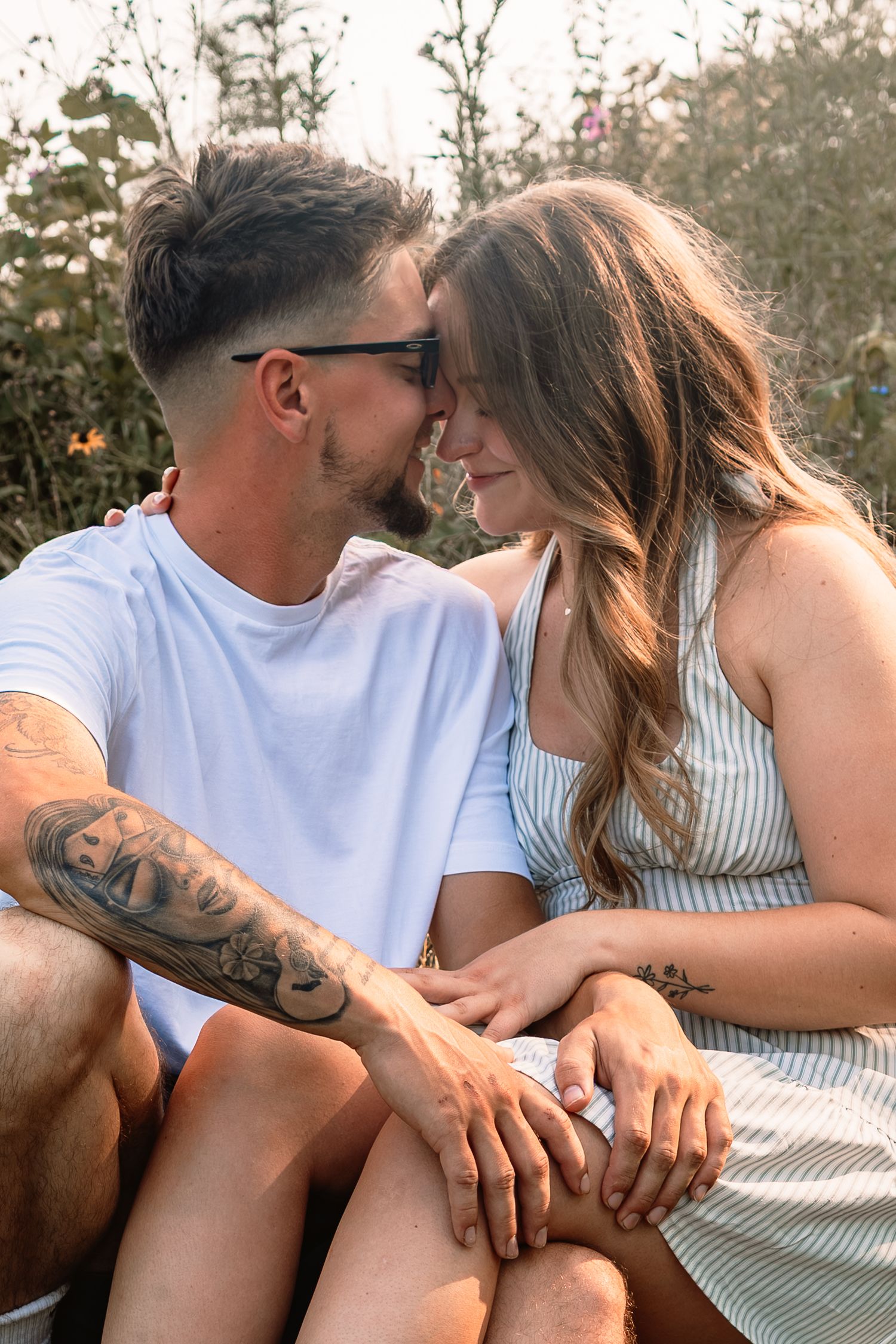 A romantic couple shares an intimate moment outdoors, with one person wearing a white t-shirt showing tattoos on their arm.