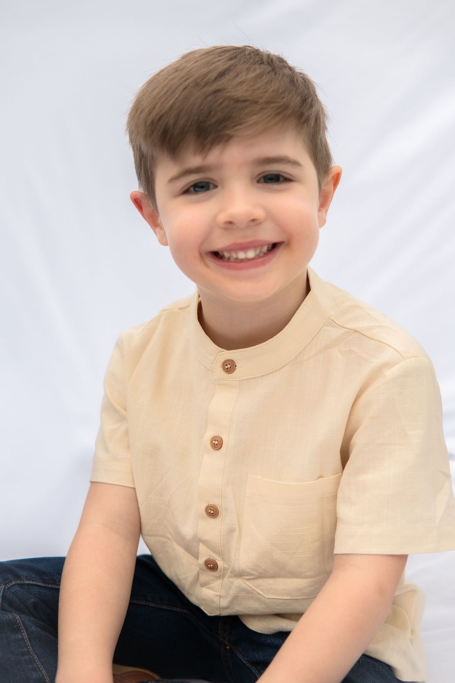 Portrait of a child wearing a tan buttoned shirt with short sleeves and blue pants against a white backdrop.
