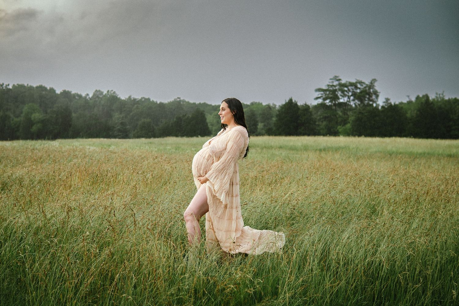 A figure in a flowing cream dress stands gracefully in a serene meadow with tall grass on a misty day.