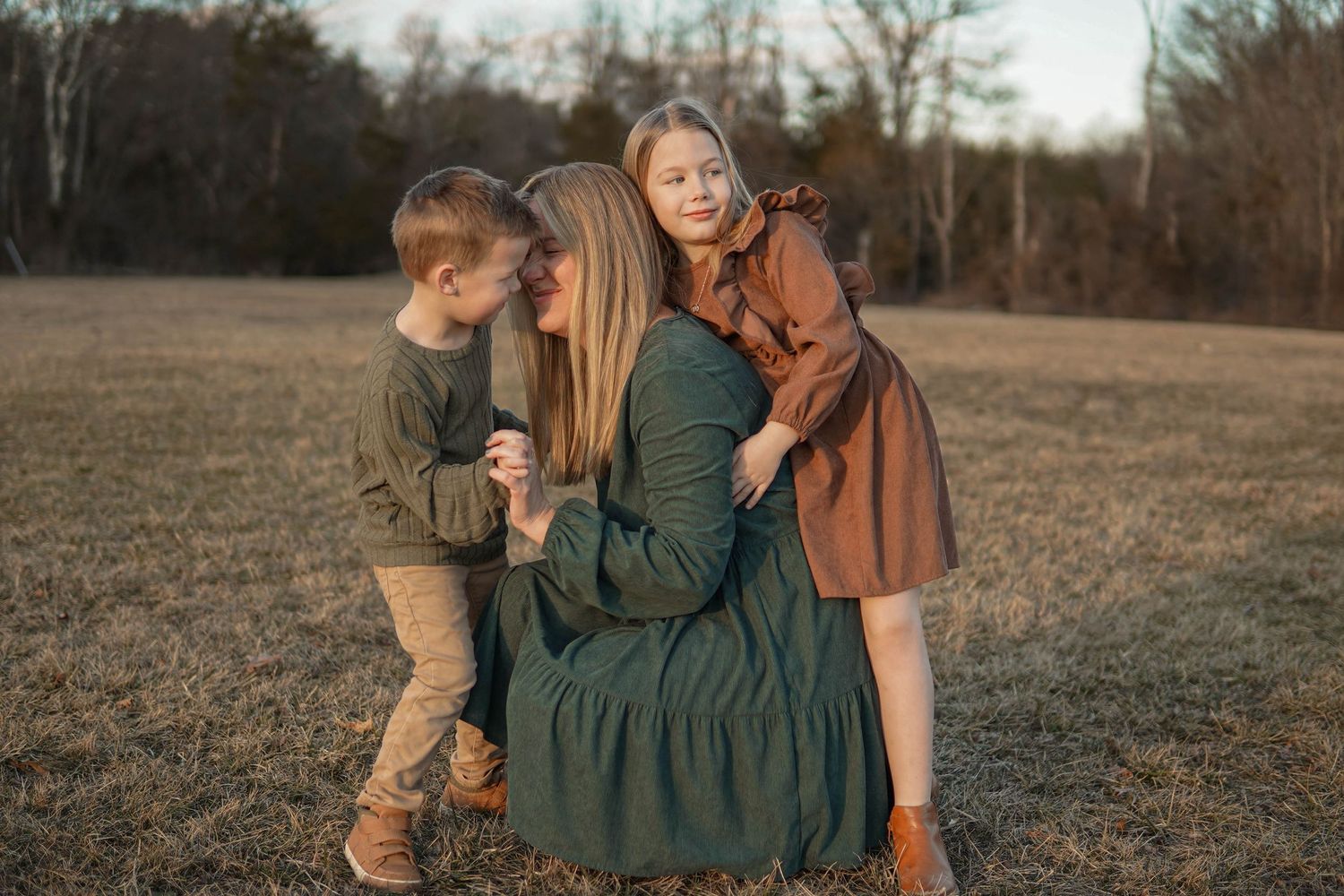 Three people in rustic autumn clothing embrace in a brown grassy field on a fall day.