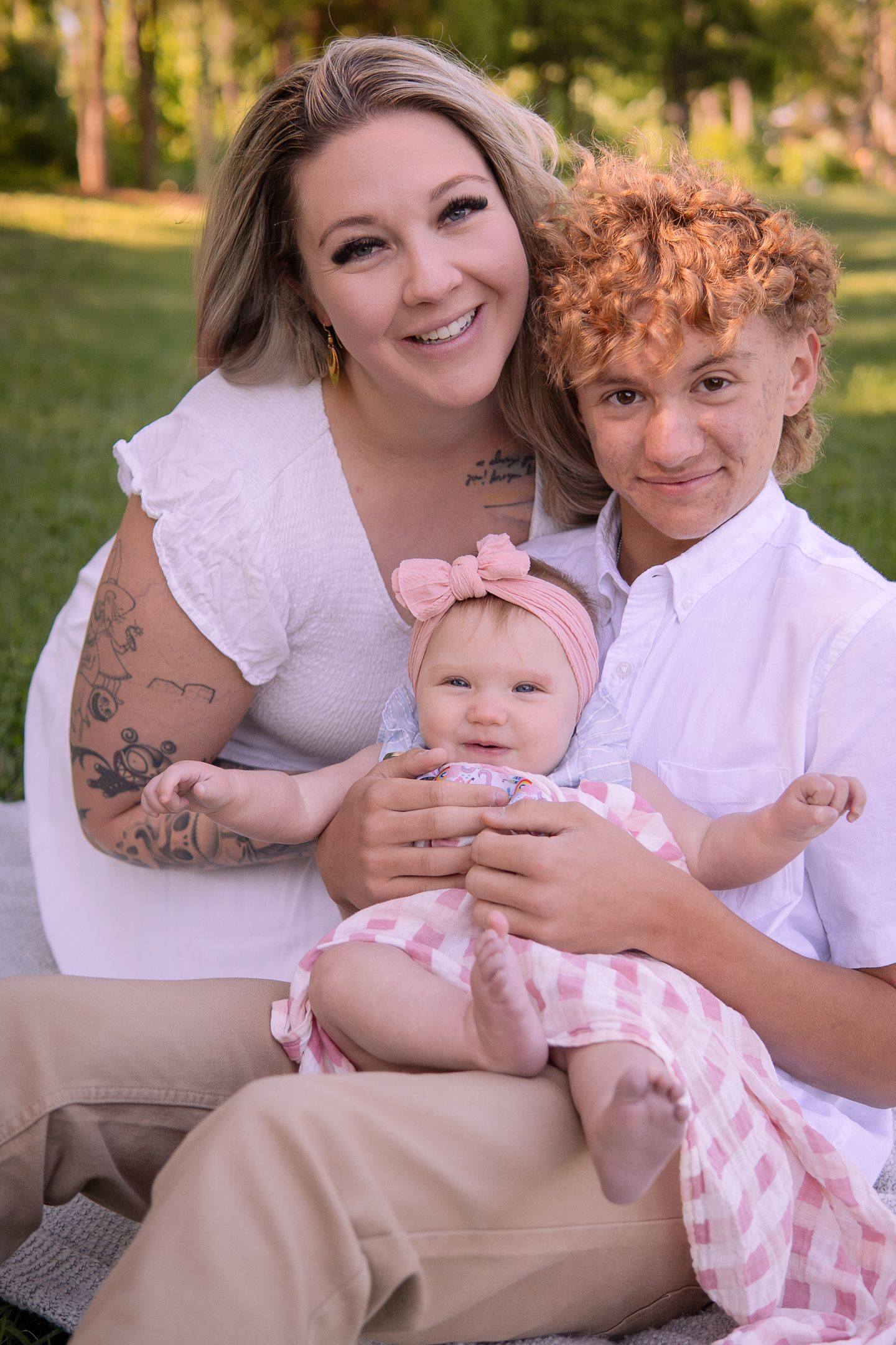 Family sitting together outdoors in white clothing with their baby wearing a pink headband and checkered outfit.
