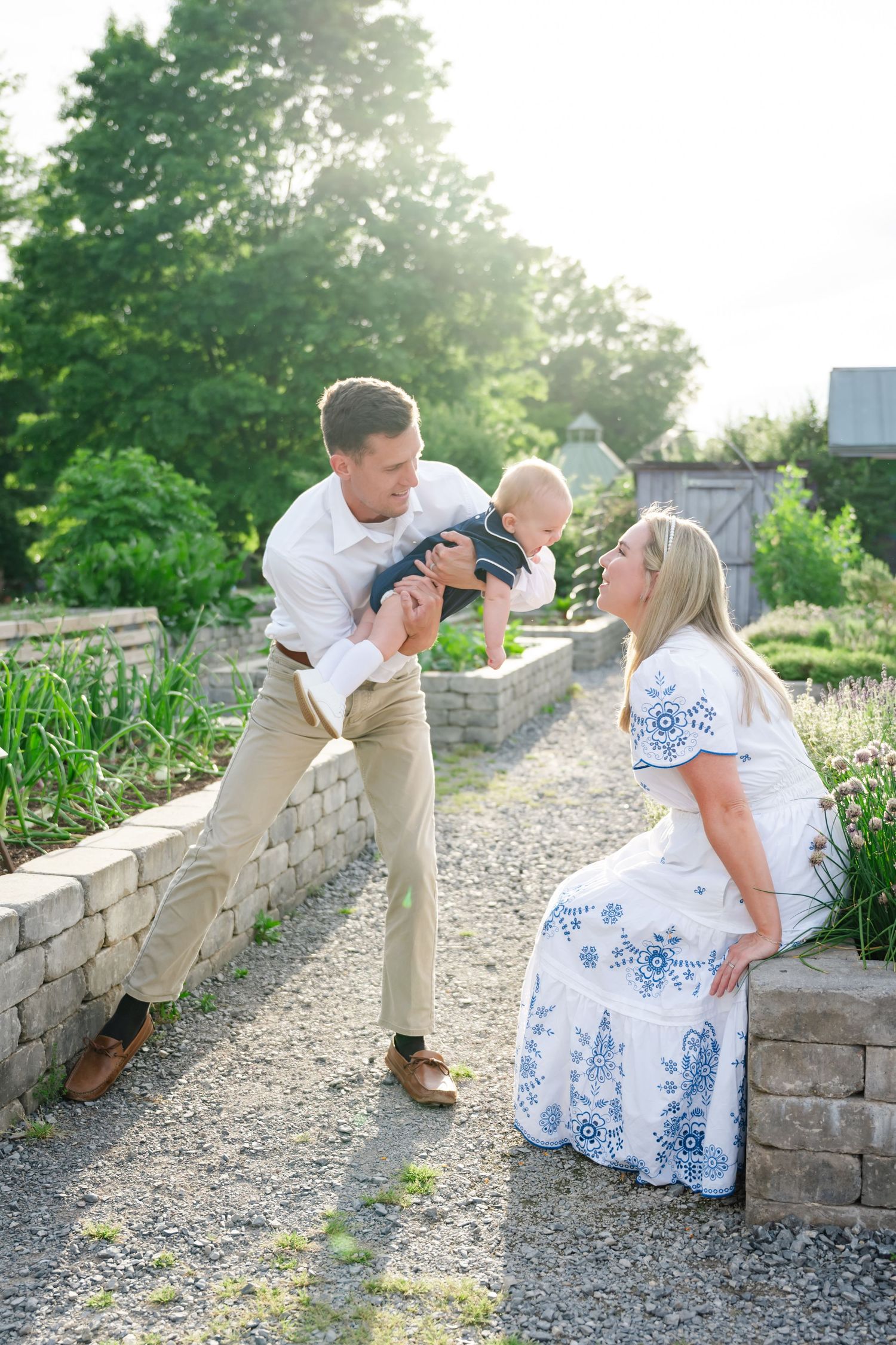 A happy family with a man lifting a baby toward a smiling woman in a white dress in a sunny garden.