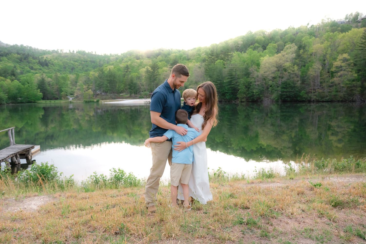 Family of three embracing by a serene lake surrounded by lush green trees during a golden hour outdoor photo session.
