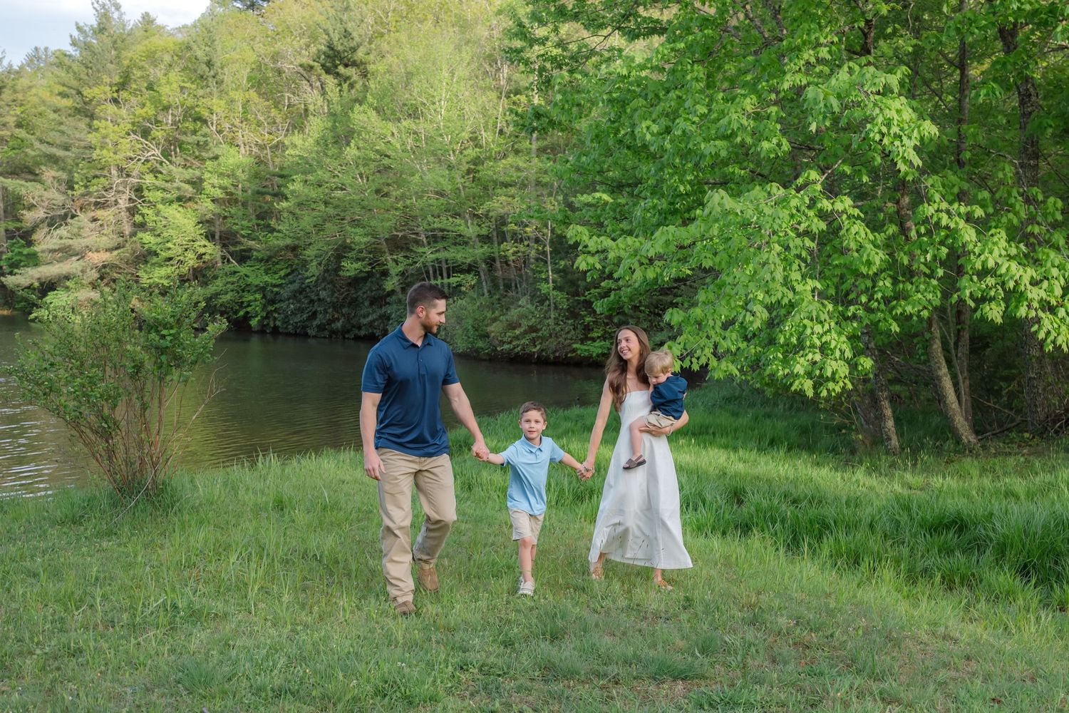 Family of four walking hand-in-hand through lush green meadow near a pond, parents holding children on a sunny day.