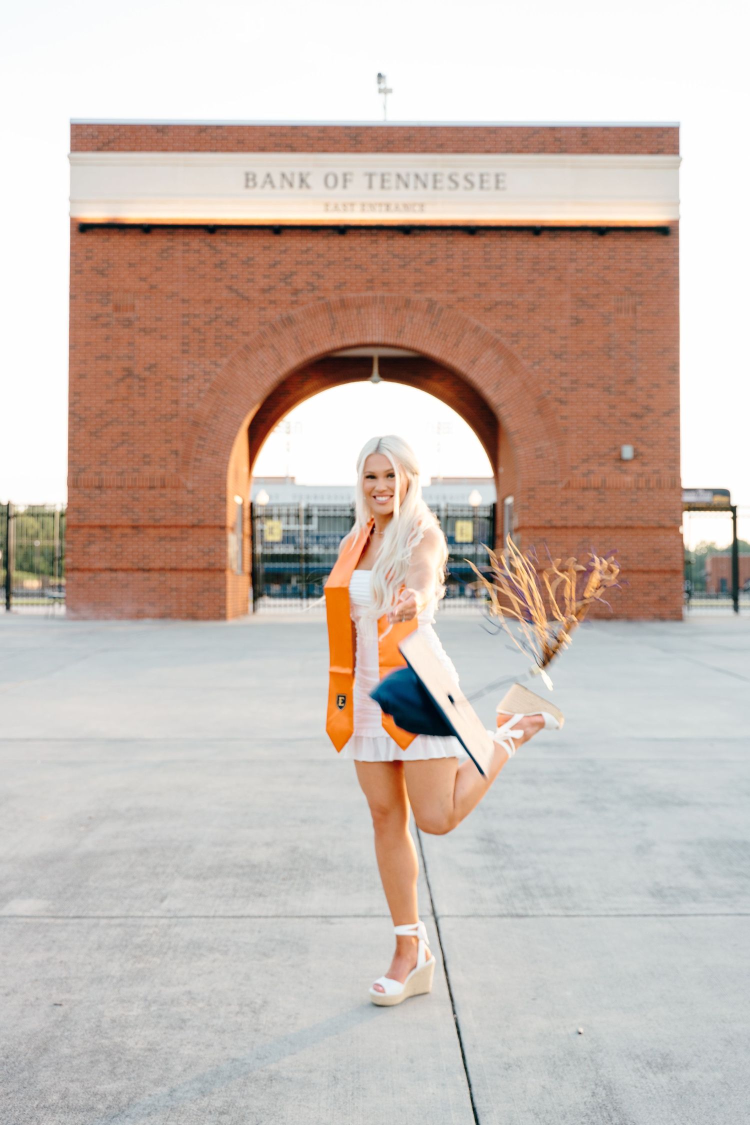 Graduate in white dress performs playful jumping poses in front of brick building.