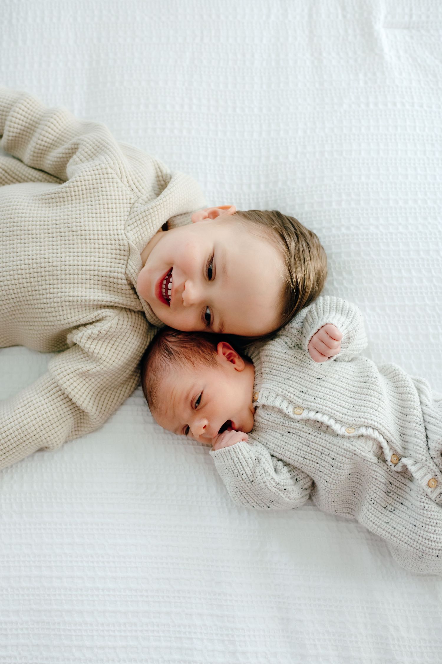 Two children in neutral sweaters laying close together on white bedding.