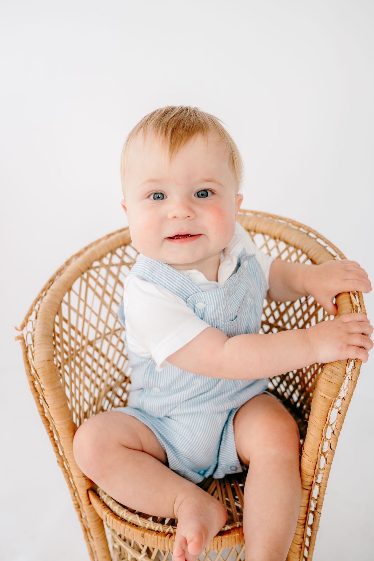 Baby wearing blue outfit sitting in wicker chair against white background.
