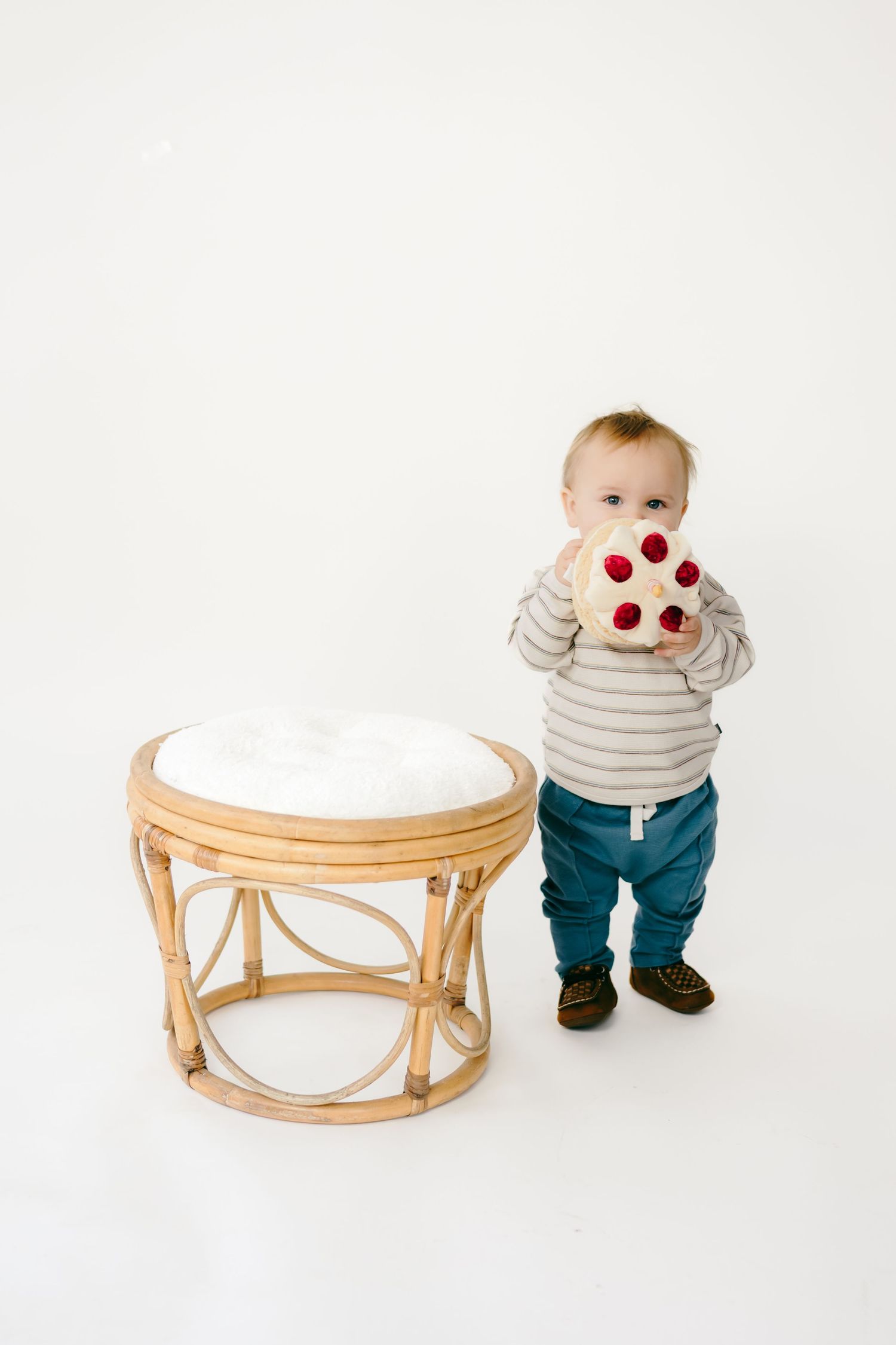 Small child in striped shirt and blue pants plays near a wooden rattan table in a minimalist setting.