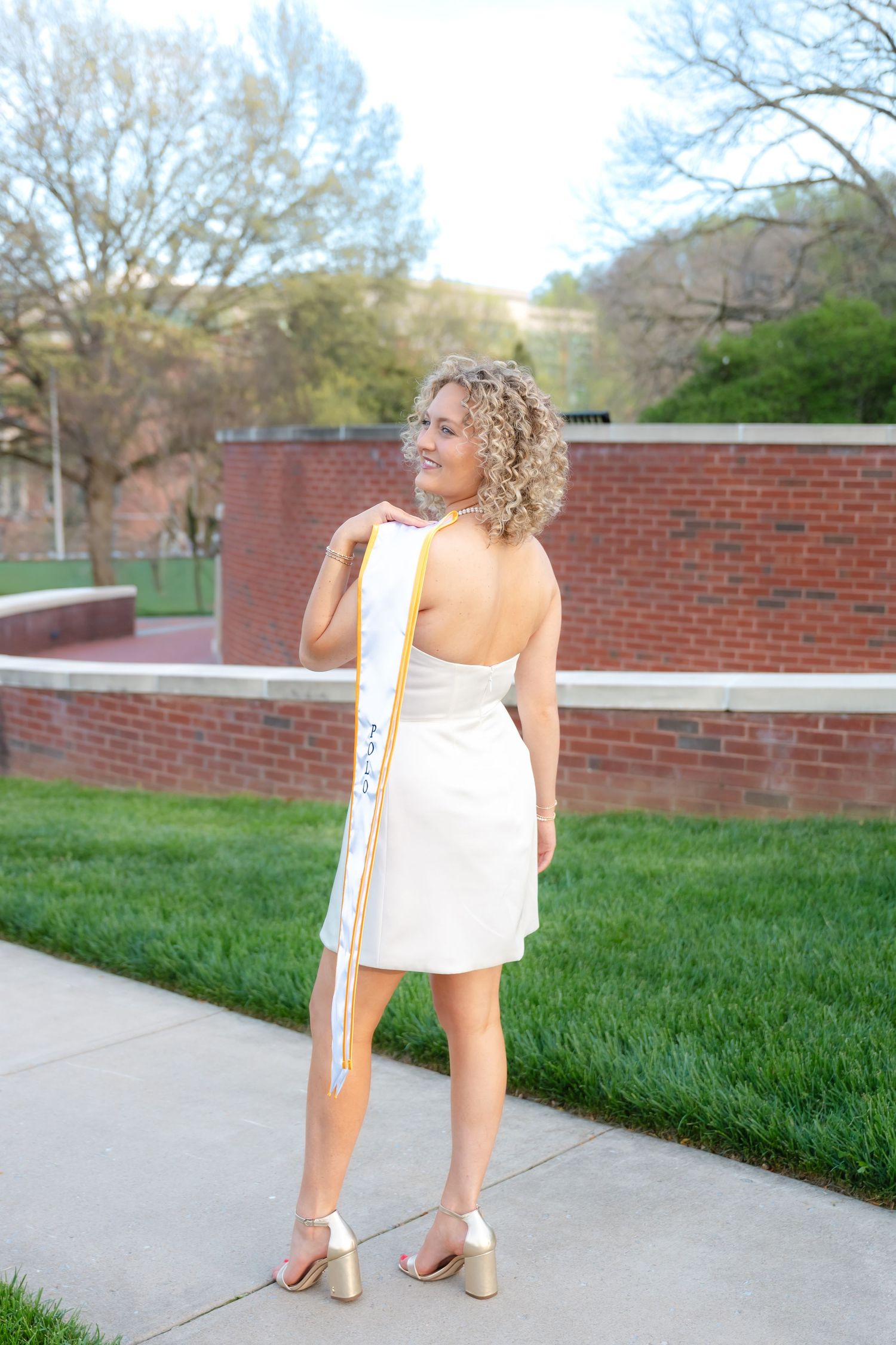 Young woman in white dress and gold sash poses outdoors on campus sidewalk with brick building and green lawn.