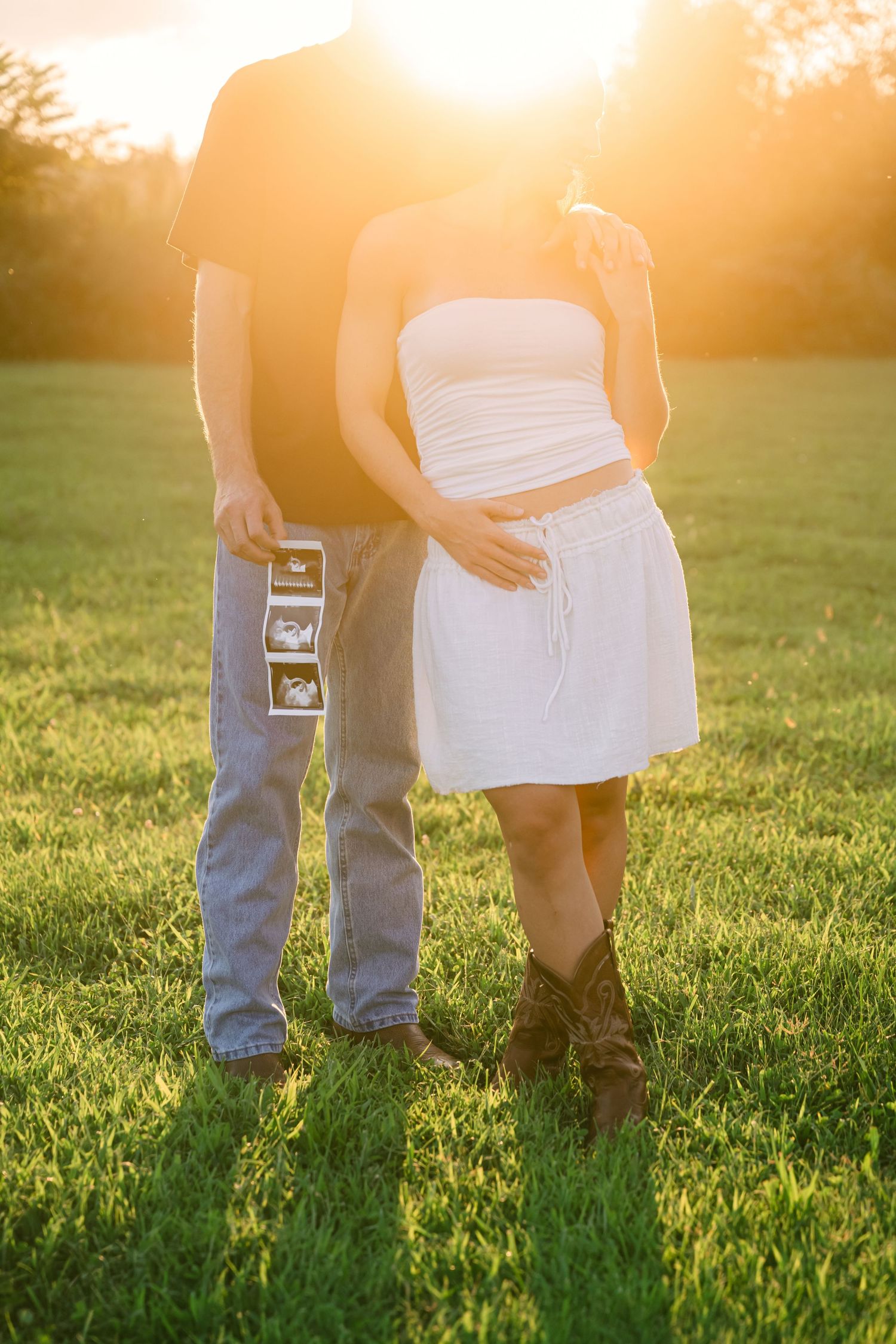 Couple holding ultrasound photos at golden hour in a field, woman in white dress touching baby bump, maternity announcement photo.