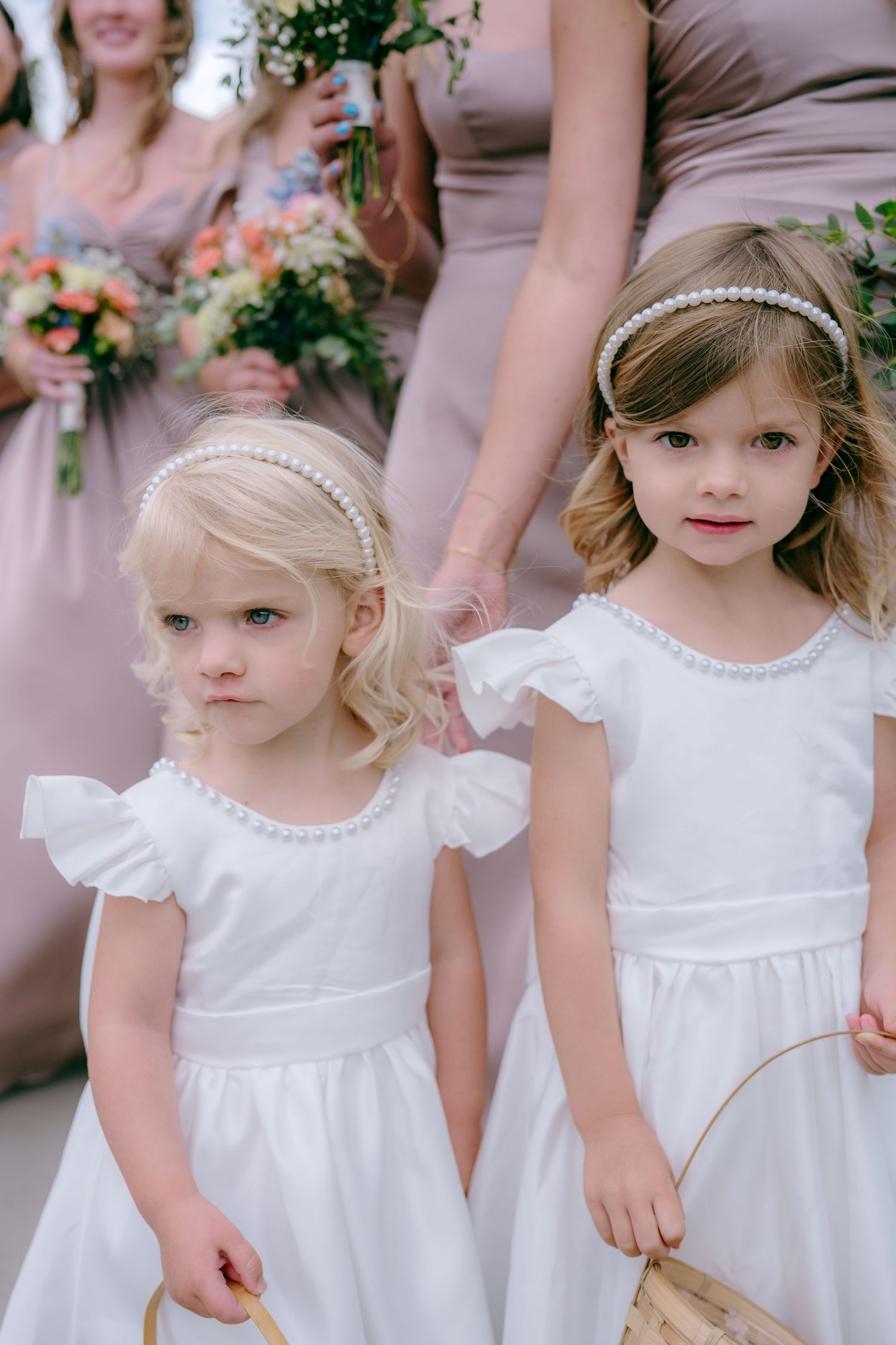 Flower girls in white dresses with pearl headbands stand together at outdoor wedding.